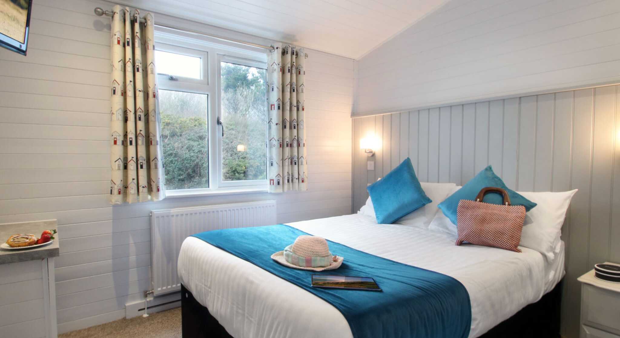 A cozy guest room featuring a double bed with white linens and turquoise accents, complemented by decorative pillows. There is a small table next to the bed with a book and a sun hat, and a window dressed with patterned curtains overlooks greenery outside. A flat-screen TV is mounted on the wall.