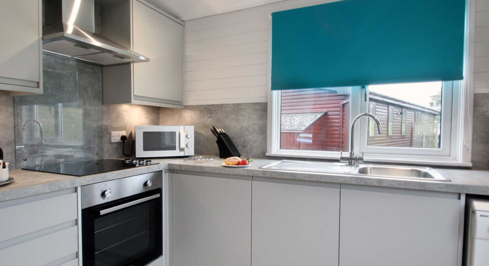 Modern kitchen featuring gray cabinets, stainless steel appliances, and a turquoise blind over the window. A countertop with a sink and microwave is visible, along with a few utensils and a plate of food.