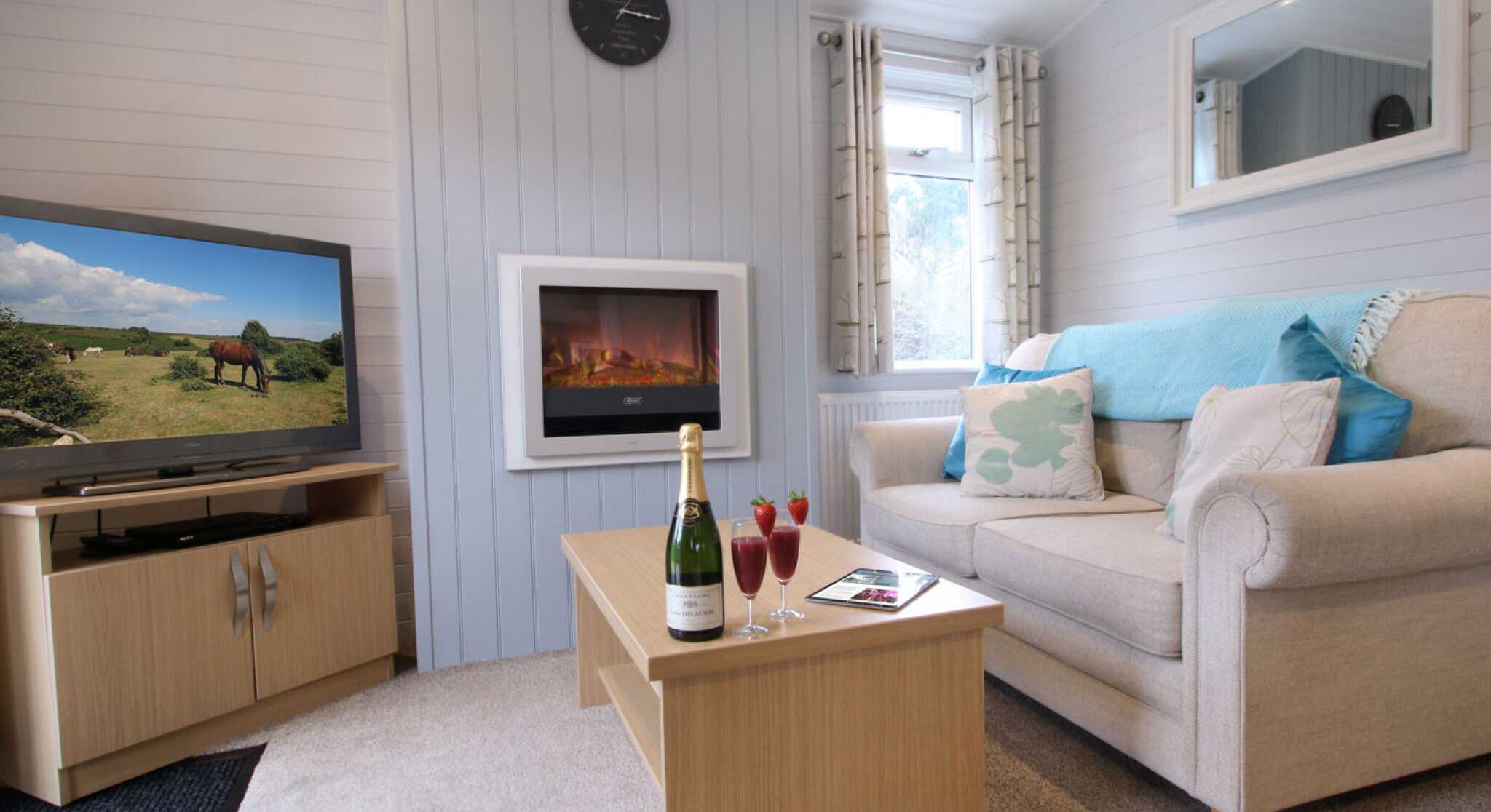 A cozy living room featuring a beige sofa with decorative pillows, a wooden coffee table with champagne and cocktails, a wall-mounted electric fireplace, and a television showing a rural landscape. Light grey paneling and natural light create a warm ambiance.