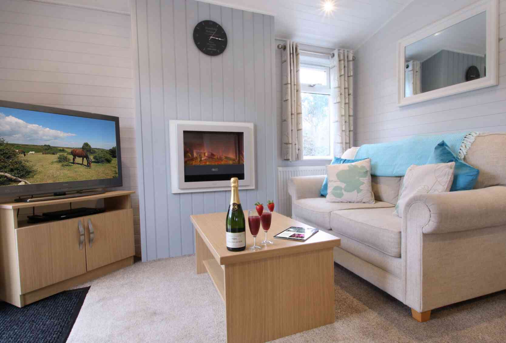 A cozy living room featuring a beige sofa with decorative pillows, a wooden coffee table with champagne and cocktails, a wall-mounted electric fireplace, and a television showing a rural landscape. Light grey paneling and natural light create a warm ambiance.