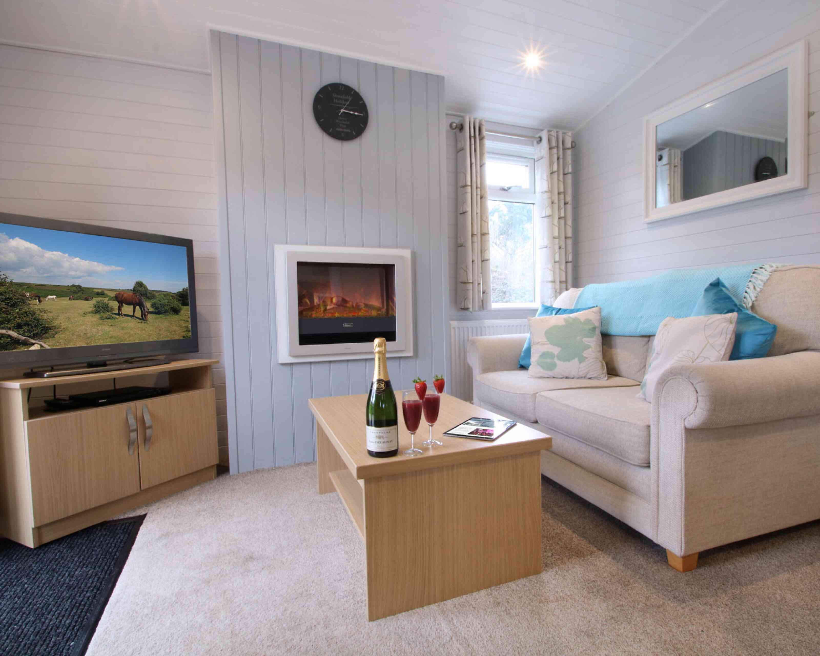 A cozy living room featuring a beige sofa with decorative pillows, a wooden coffee table with champagne and cocktails, a wall-mounted electric fireplace, and a television showing a rural landscape. Light grey paneling and natural light create a warm ambiance.