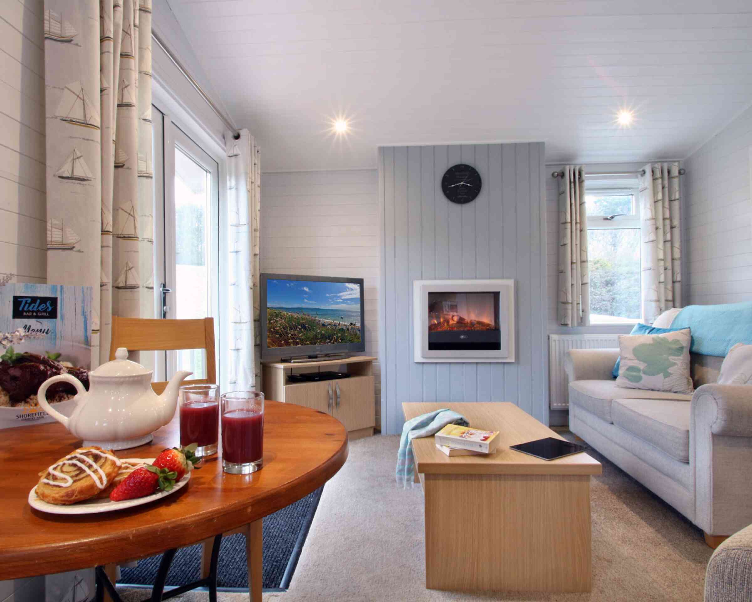 A cozy living room with light walls and a large window. A comfortable sofa is paired with a coffee table, while a dining table holds a teapot and glasses of juice, alongside pastries and strawberries. A TV is visible on one side, and a modern fireplace adds warmth to the setting. Natural light fills the space, enhancing its inviting atmosphere.