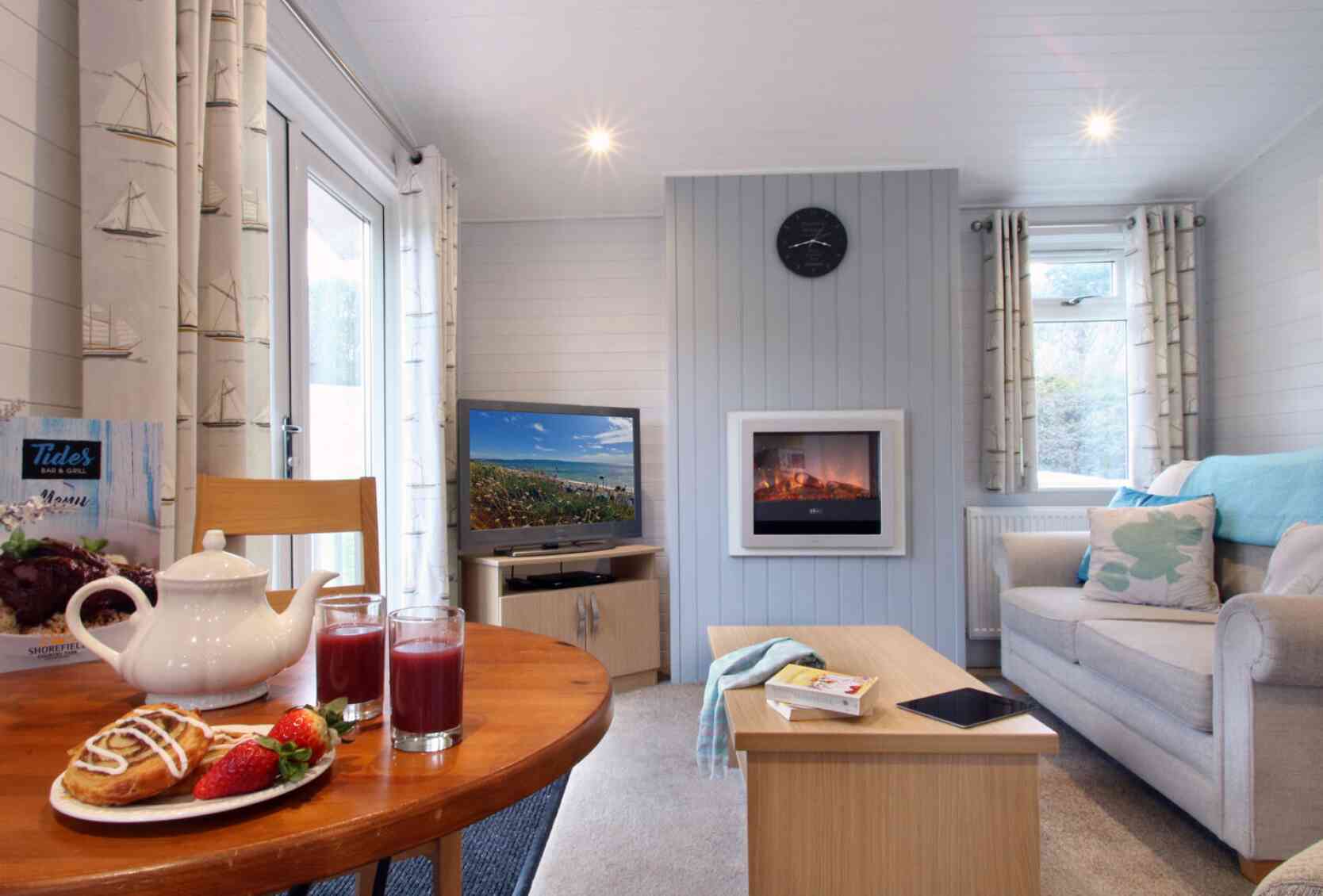 A cozy living room with light walls and a large window. A comfortable sofa is paired with a coffee table, while a dining table holds a teapot and glasses of juice, alongside pastries and strawberries. A TV is visible on one side, and a modern fireplace adds warmth to the setting. Natural light fills the space, enhancing its inviting atmosphere.