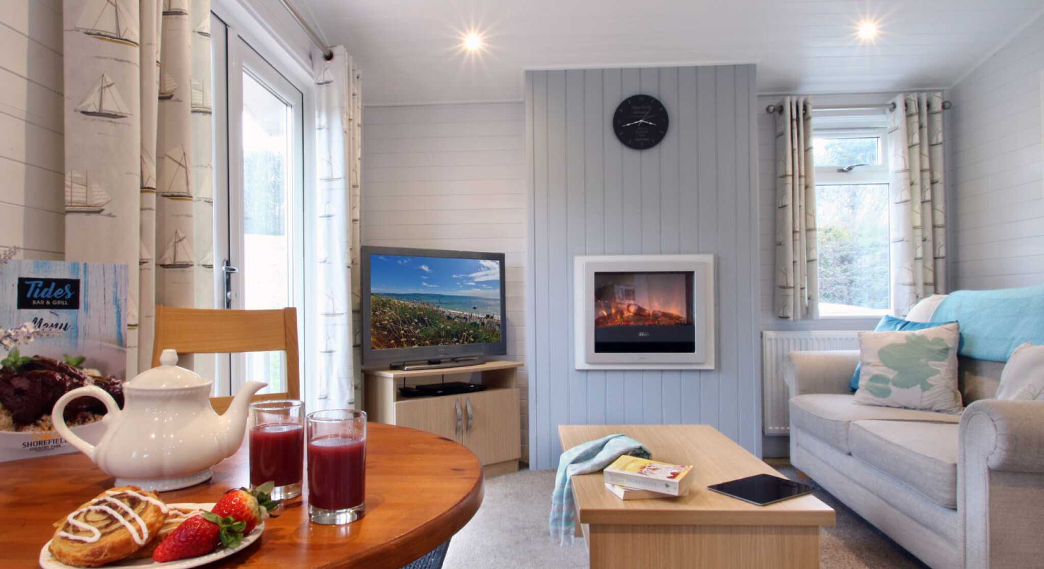 A cozy living room with light walls and a large window. A comfortable sofa is paired with a coffee table, while a dining table holds a teapot and glasses of juice, alongside pastries and strawberries. A TV is visible on one side, and a modern fireplace adds warmth to the setting. Natural light fills the space, enhancing its inviting atmosphere.