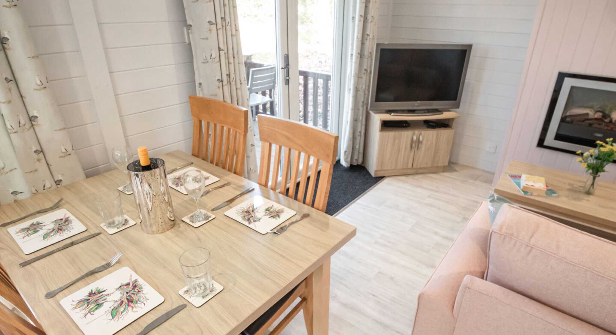 A cozy dining area features a wooden table set with decorative plates and glasses. In the background, a living space includes a pink sofa, a television on a wooden stand, and a bouquet of flowers on a side table. Large windows offer natural light and a view of a deck outside.