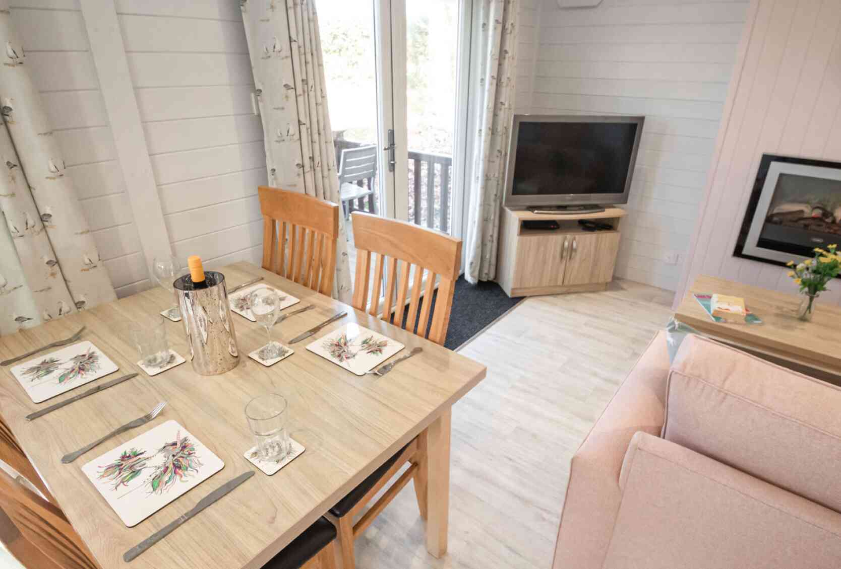A cozy dining area features a wooden table set with decorative plates and glasses. In the background, a living space includes a pink sofa, a television on a wooden stand, and a bouquet of flowers on a side table. Large windows offer natural light and a view of a deck outside.