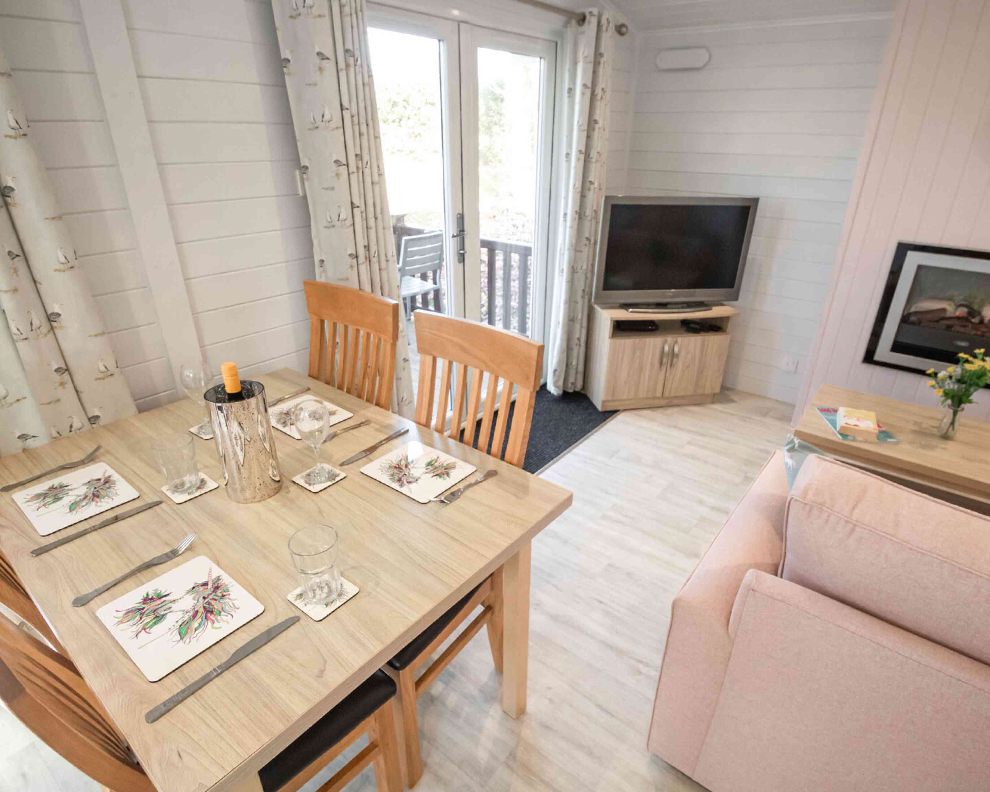 A cozy dining area features a wooden table set with decorative plates and glasses. In the background, a living space includes a pink sofa, a television on a wooden stand, and a bouquet of flowers on a side table. Large windows offer natural light and a view of a deck outside.
