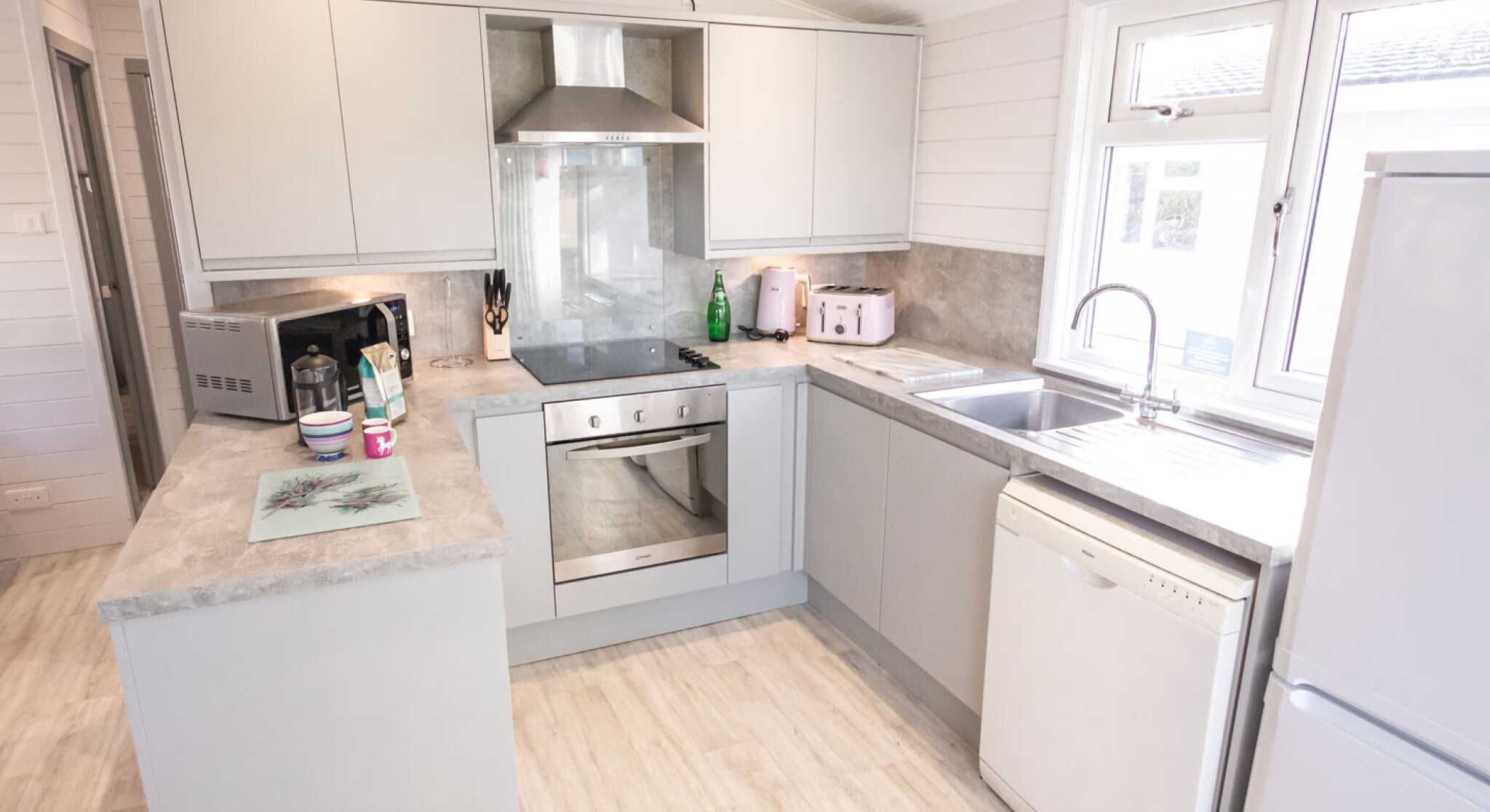 A modern kitchen featuring light gray cabinets, a stainless steel oven, and a white dishwasher. The countertop is a light gray stone, and there is a window above the sink providing natural light. Appliances include a microwave and a kettle, with a small decorative tray on the counter.