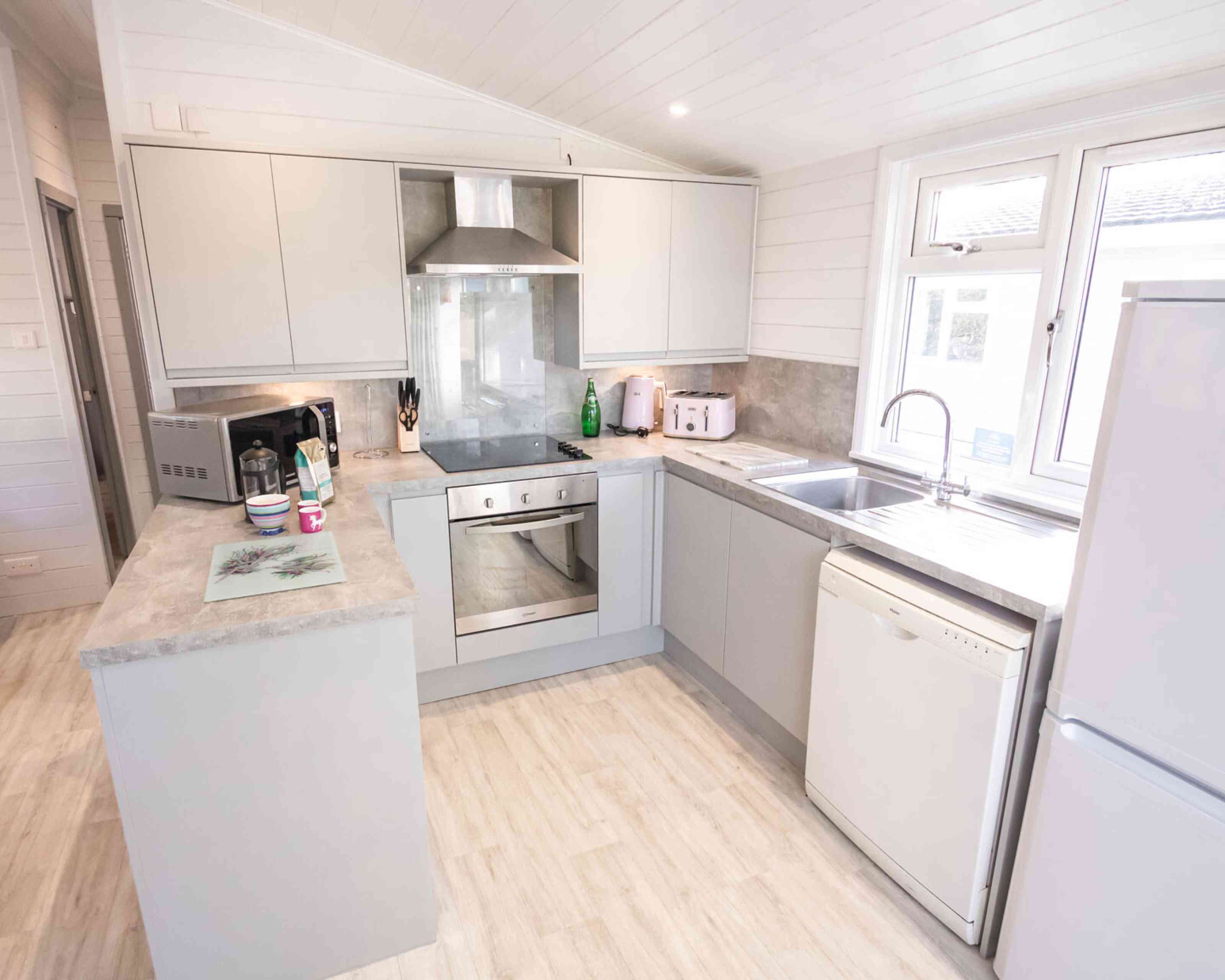 A modern kitchen featuring light gray cabinets, a stainless steel oven, and a white dishwasher. The countertop is a light gray stone, and there is a window above the sink providing natural light. Appliances include a microwave and a kettle, with a small decorative tray on the counter.