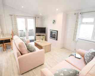 A cozy living room featuring light pink sofas with decorative pillows, a coffee table, and a mounted flat-screen TV. Large windows allow natural light, and there is a fireplace on one wall. A dining area is visible in the background, with a wooden table.