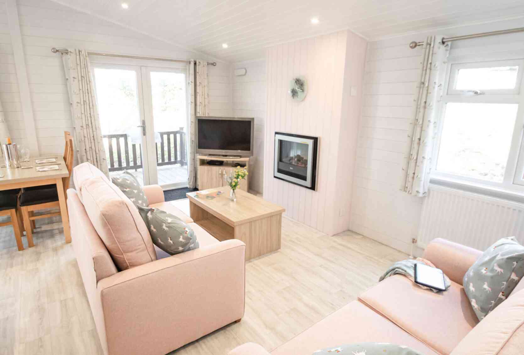 A cozy living room featuring light pink sofas with decorative pillows, a coffee table, and a mounted flat-screen TV. Large windows allow natural light, and there is a fireplace on one wall. A dining area is visible in the background, with a wooden table.