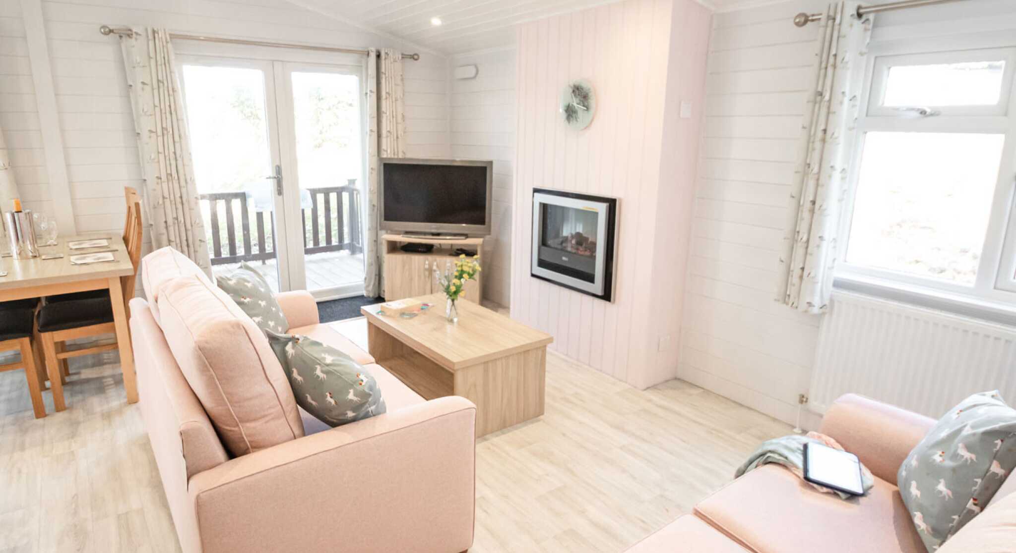 A cozy living room featuring light pink sofas with decorative pillows, a coffee table, and a mounted flat-screen TV. Large windows allow natural light, and there is a fireplace on one wall. A dining area is visible in the background, with a wooden table.