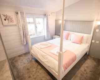 A cozy bedroom featuring a four-poster bed with white bedding and soft pink accents. Large windows let in natural light, complemented by light-colored walls and decorative curtains. A small nightstand is positioned beside the bed, and a plush carpet adds warmth to the space.