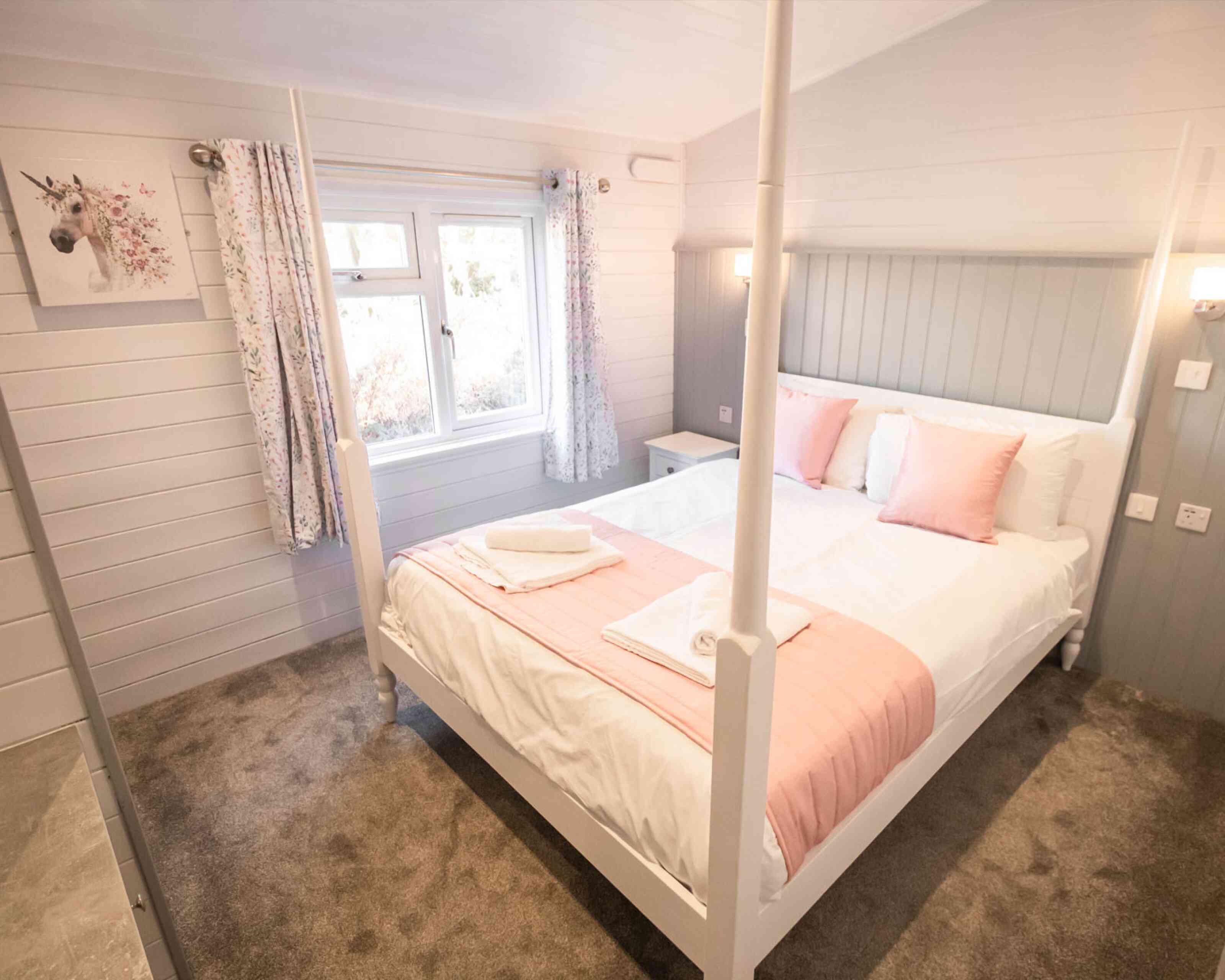 A cozy bedroom featuring a four-poster bed with white bedding and soft pink accents. Large windows let in natural light, complemented by light-colored walls and decorative curtains. A small nightstand is positioned beside the bed, and a plush carpet adds warmth to the space.