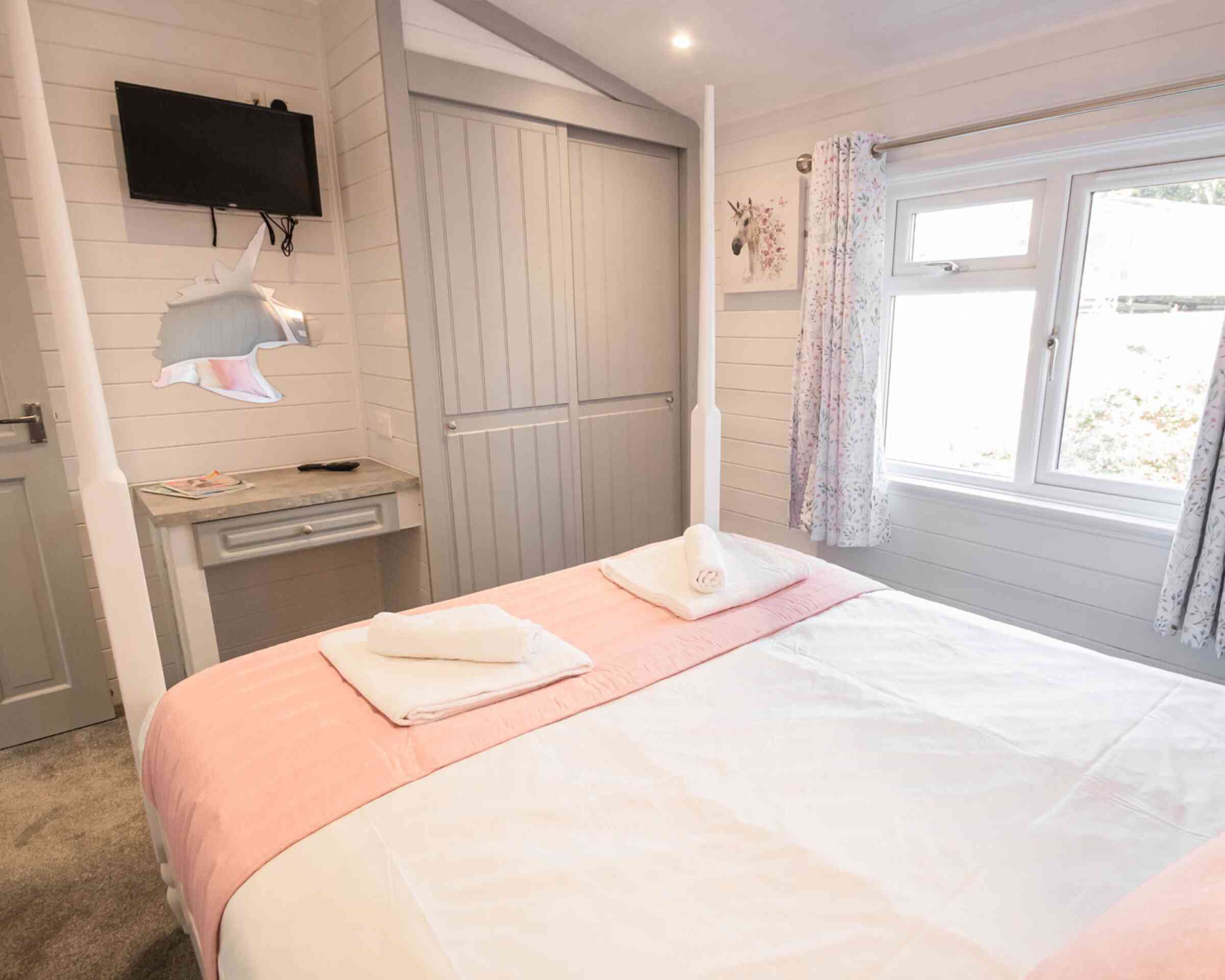 A cozy bedroom featuring a double bed with pink bedding and neatly rolled towels. The room includes a wall-mounted TV, a small desk with a lamp, and large windows with floral curtains. The walls have a light wood paneling for a warm, inviting atmosphere.