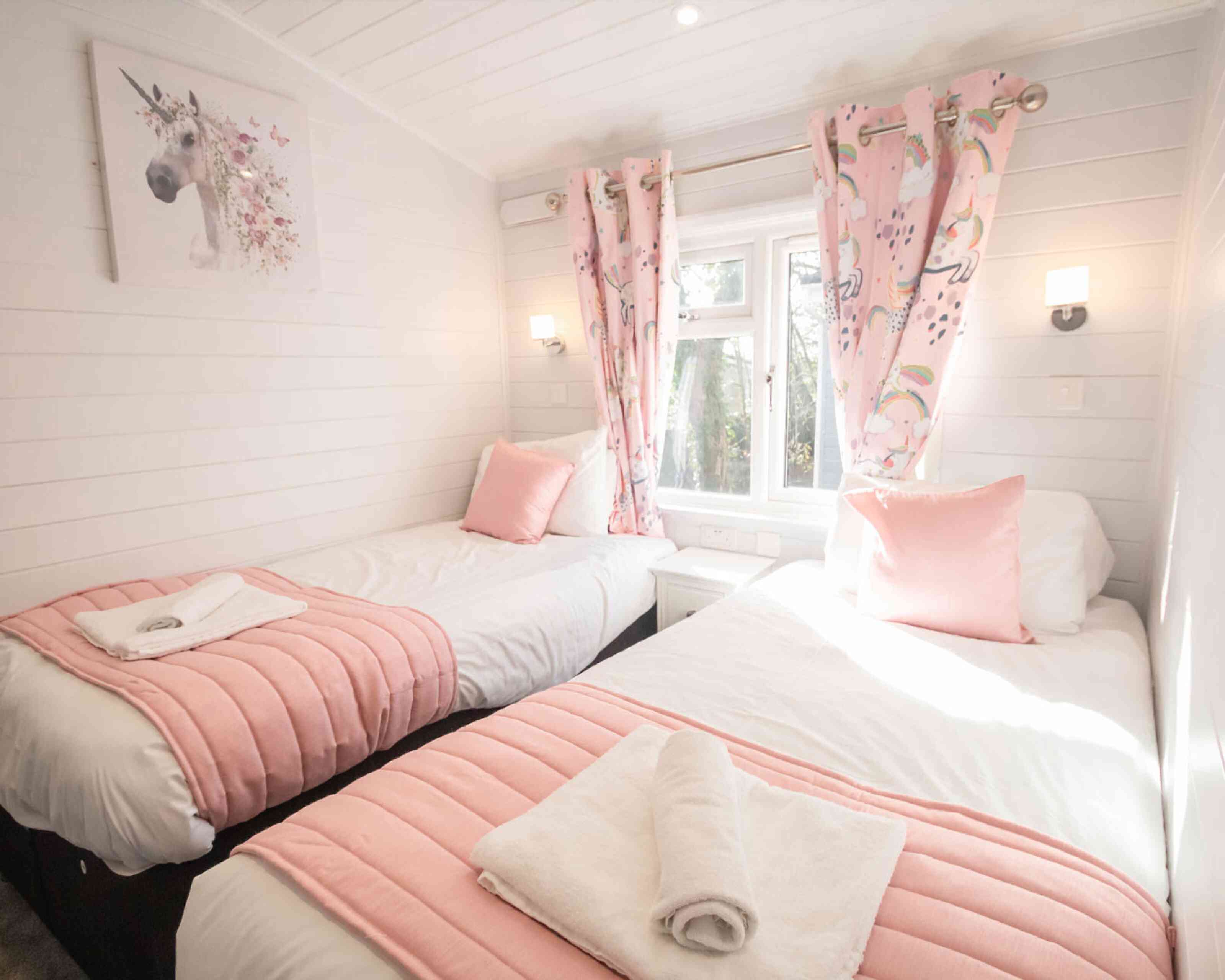 A cozy bedroom with two twin beds dressed in white linens and pink bedspreads. The room features light wood paneling and bright natural light streaming through a window adorned with pink curtains decorated with unicorns. There is a unicorn-themed artwork on the wall and soft pink pillows on the beds. A towel is neatly placed on each bed.