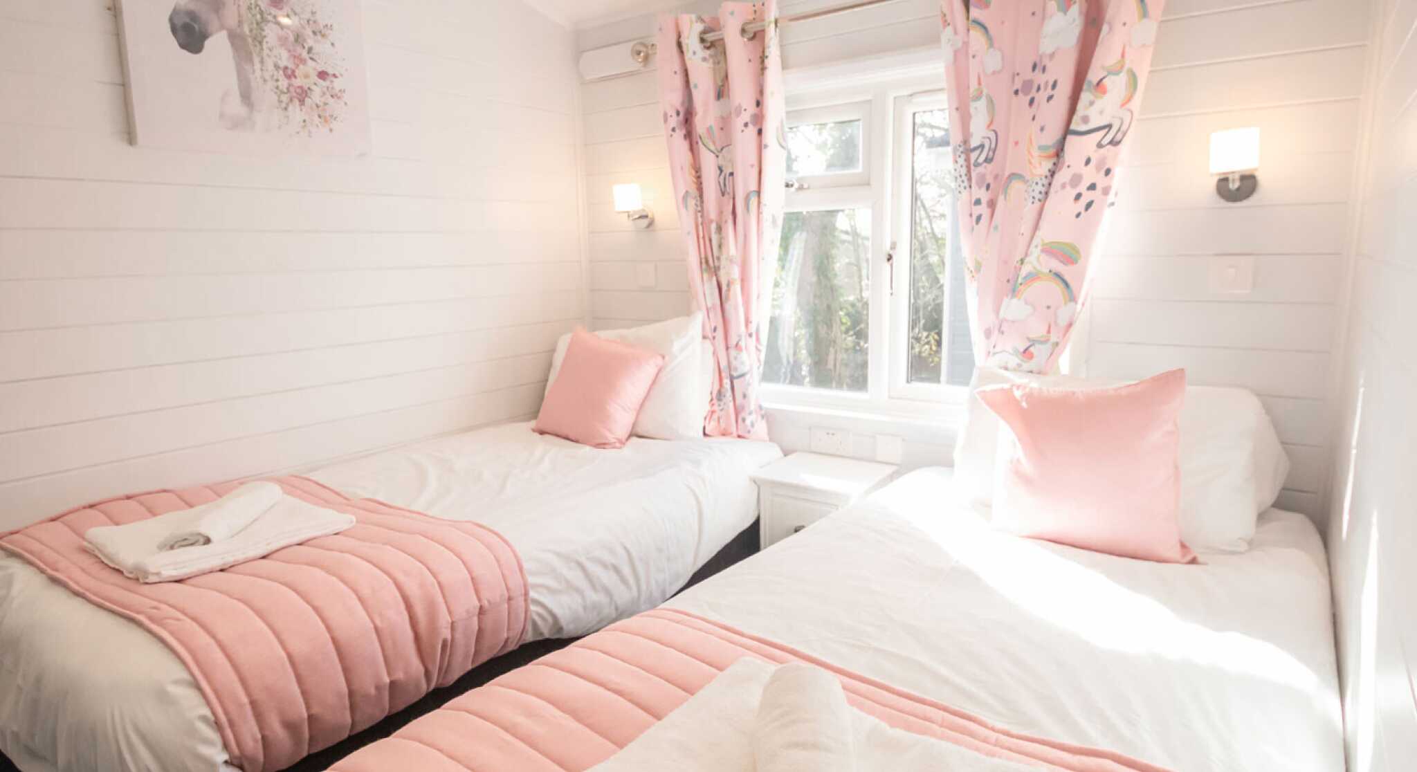 A cozy bedroom with two twin beds dressed in white linens and pink bedspreads. The room features light wood paneling and bright natural light streaming through a window adorned with pink curtains decorated with unicorns. There is a unicorn-themed artwork on the wall and soft pink pillows on the beds. A towel is neatly placed on each bed.