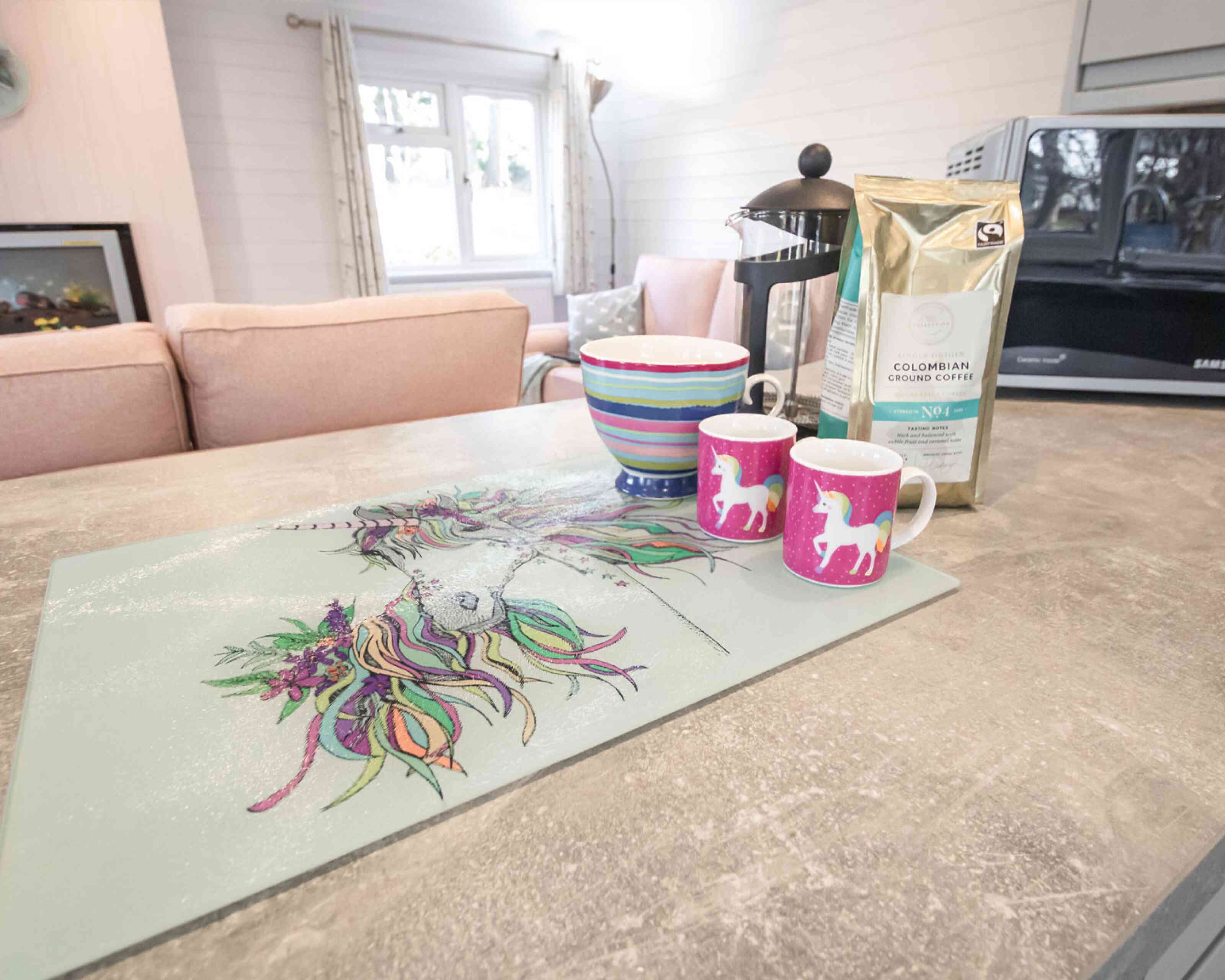 A colorful kitchen countertop featuring a unicorn-themed placemat, two pink unicorn mugs, a striped bowl, and a bag of Colombian coffee. In the background, there are soft pink sofas and a microwave.
