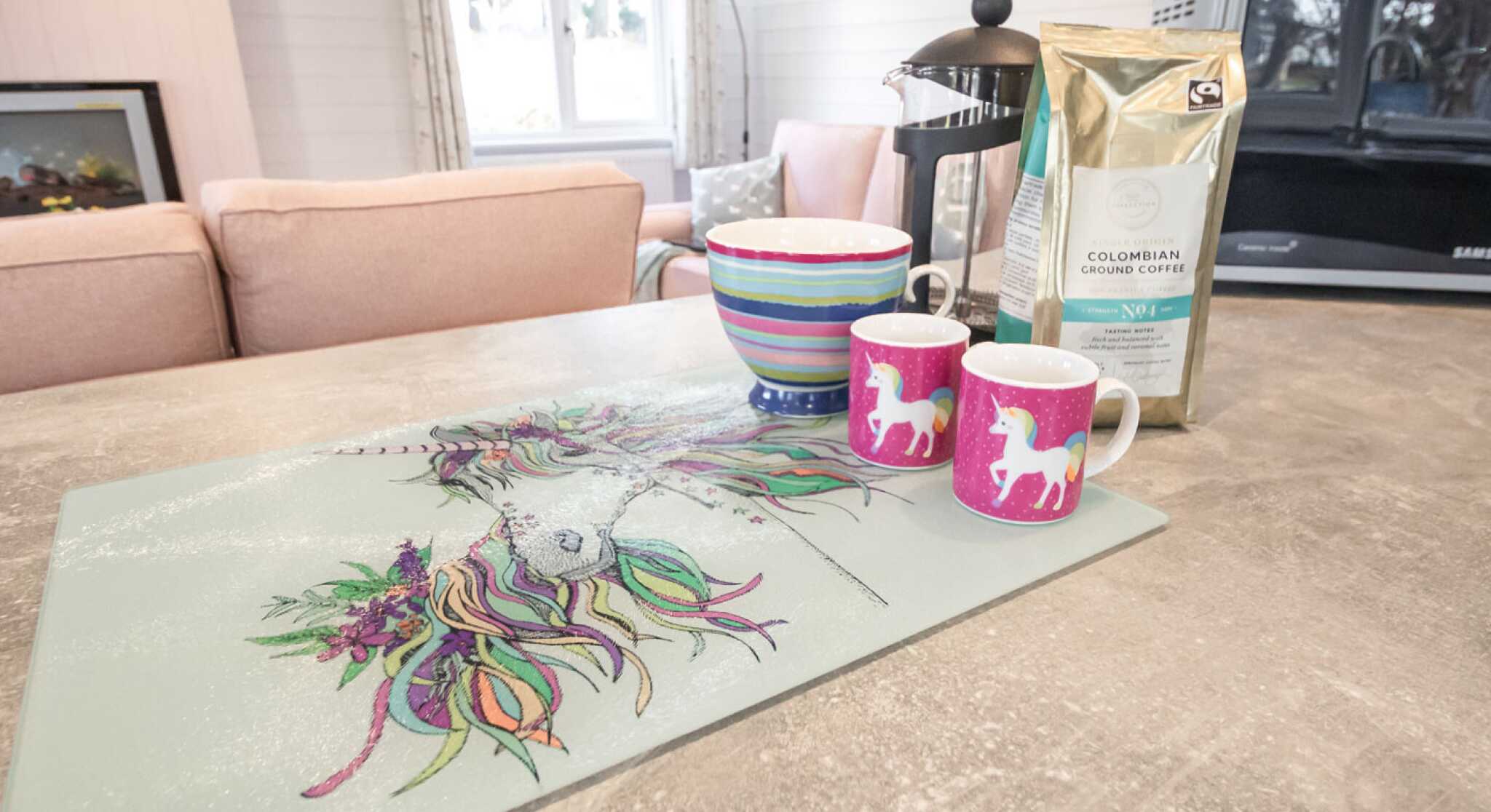 A colorful kitchen countertop featuring a unicorn-themed placemat, two pink unicorn mugs, a striped bowl, and a bag of Colombian coffee. In the background, there are soft pink sofas and a microwave.