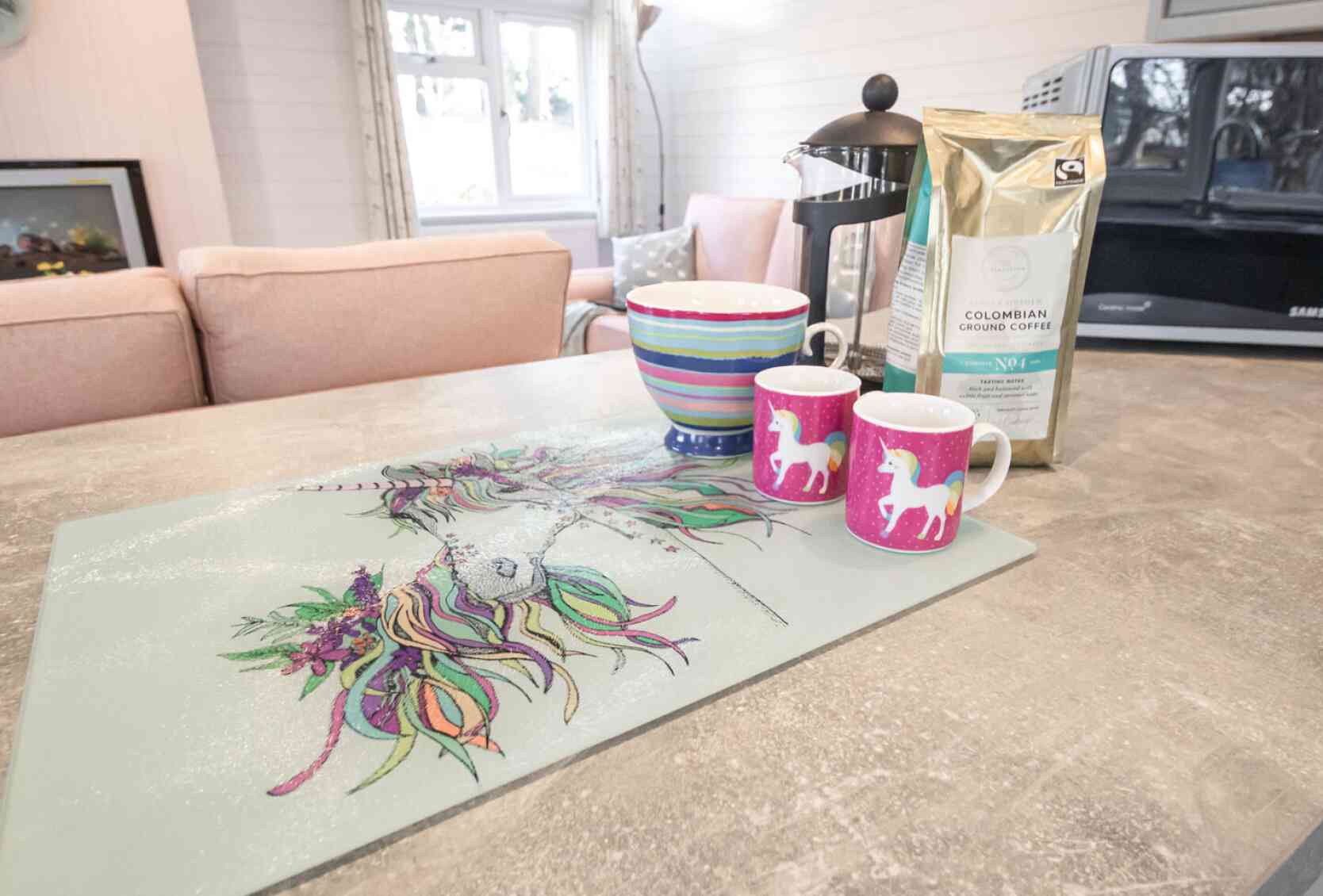 A colorful kitchen countertop featuring a unicorn-themed placemat, two pink unicorn mugs, a striped bowl, and a bag of Colombian coffee. In the background, there are soft pink sofas and a microwave.