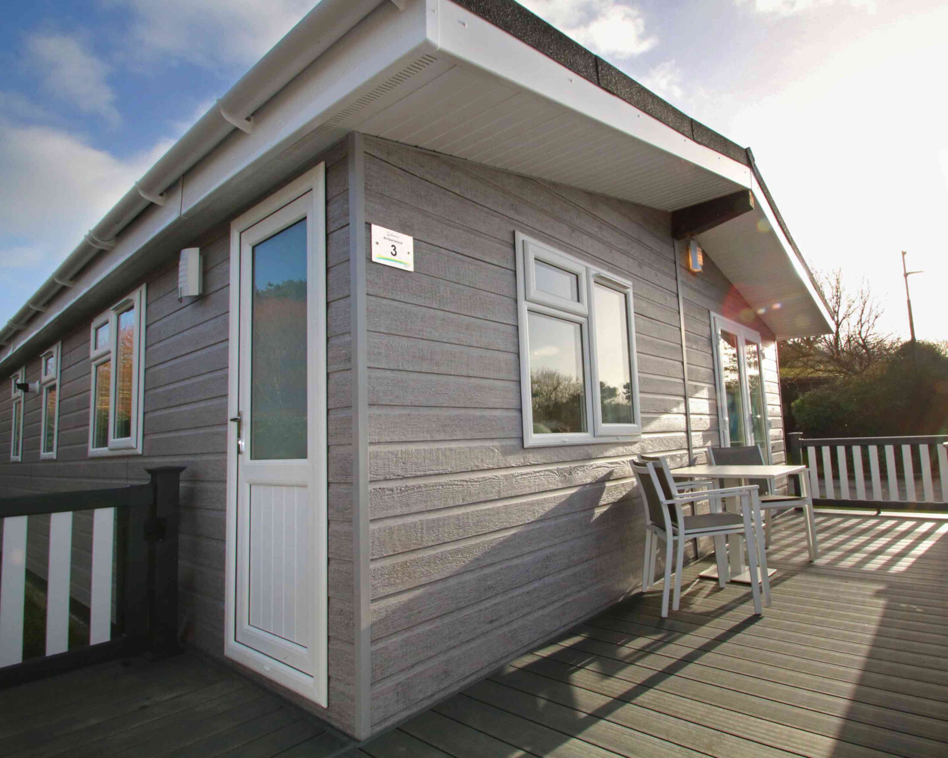 A modern wooden cabin with gray siding, featuring a white door and large windows. A small table and chairs are on the deck, surrounded by greenery under a clear blue sky.