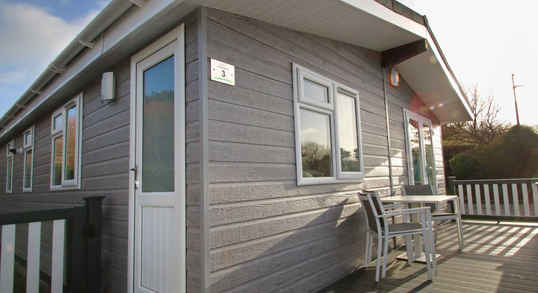 A modern wooden cabin with gray siding, featuring a white door and large windows. A small table and chairs are on the deck, surrounded by greenery under a clear blue sky.