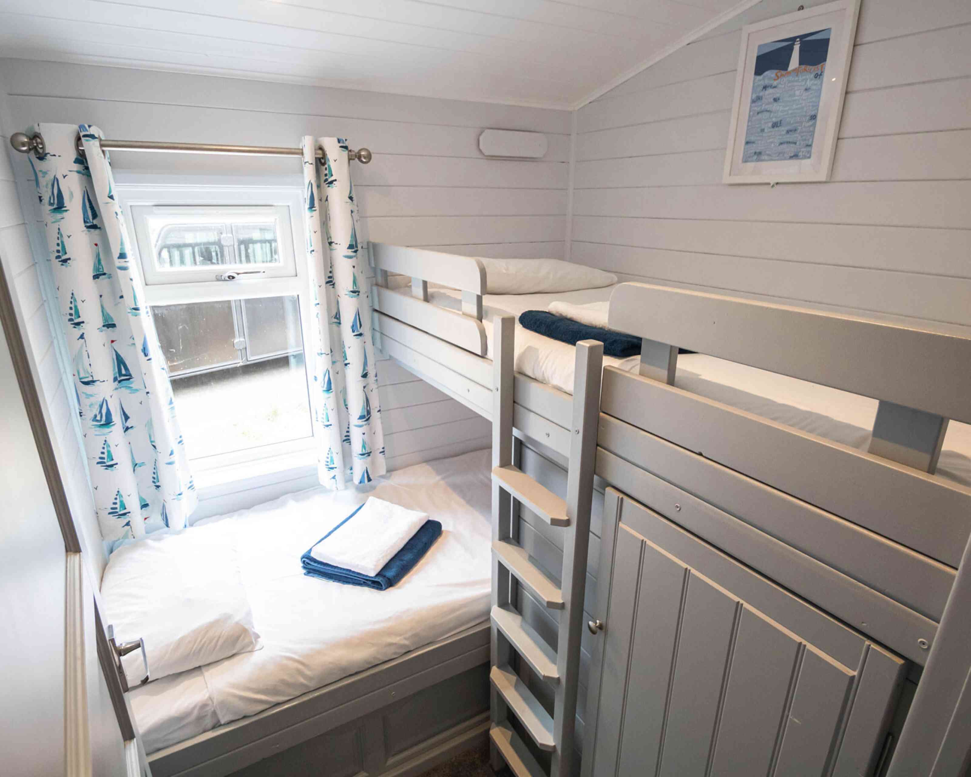A cozy bedroom with a bunk bed and white walls. The upper bunk has a soft blanket and pillow, while the lower bed is neatly made with towels. Light blue curtains with sailboat patterns frame a window that lets in natural light. A ladder leads to the upper bunk, and a white framed picture hangs on the wall.