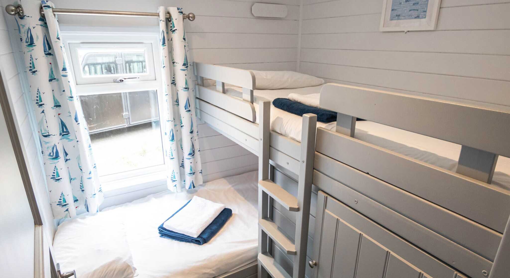 A cozy bedroom with a bunk bed and white walls. The upper bunk has a soft blanket and pillow, while the lower bed is neatly made with towels. Light blue curtains with sailboat patterns frame a window that lets in natural light. A ladder leads to the upper bunk, and a white framed picture hangs on the wall.