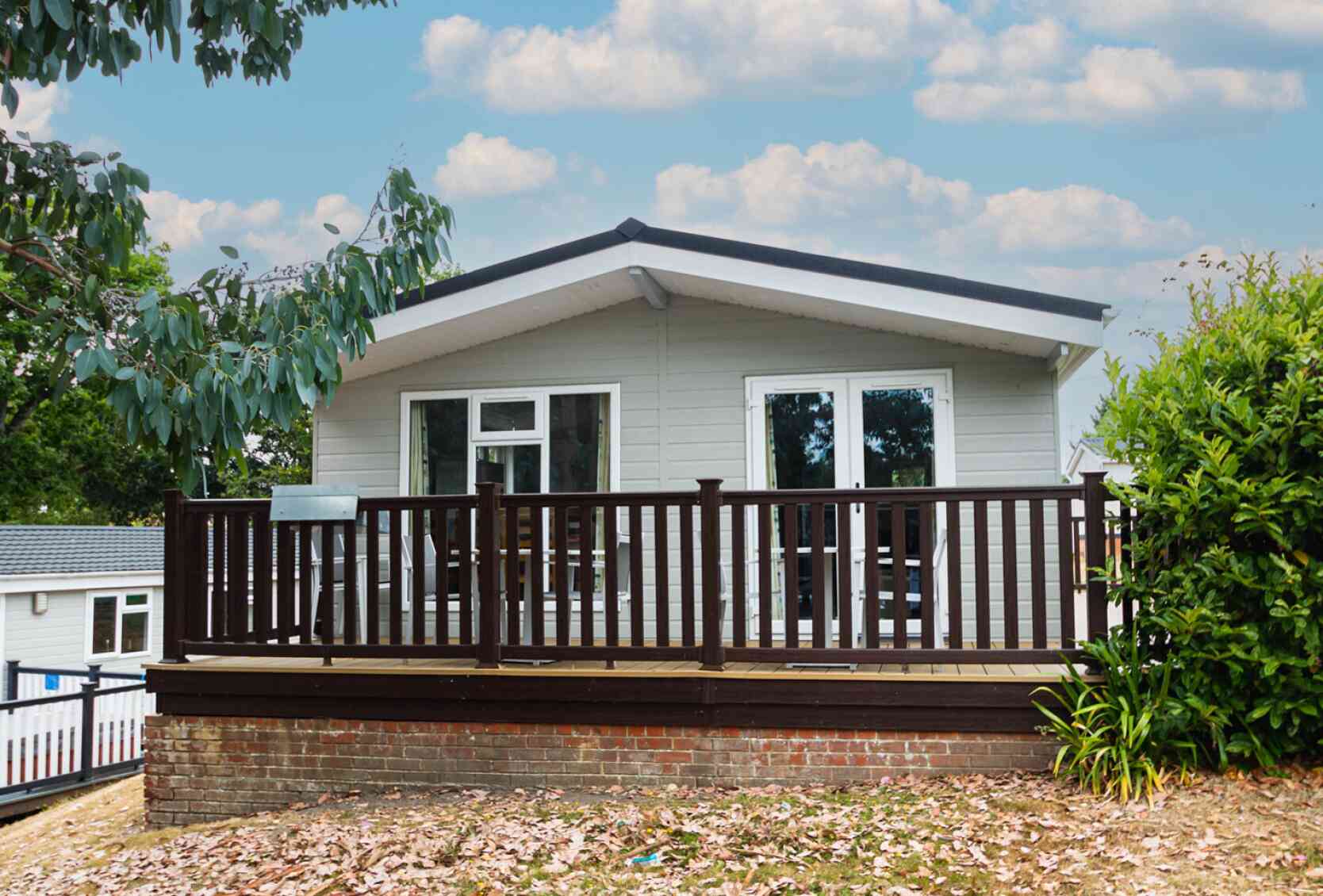 A modern single-story house with a sloped roof, featuring a wooden deck surrounded by greenery and pastel blue skies with fluffy clouds in the background.