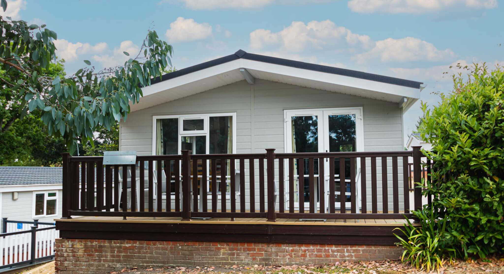 A modern single-story house with a sloped roof, featuring a wooden deck surrounded by greenery and pastel blue skies with fluffy clouds in the background.