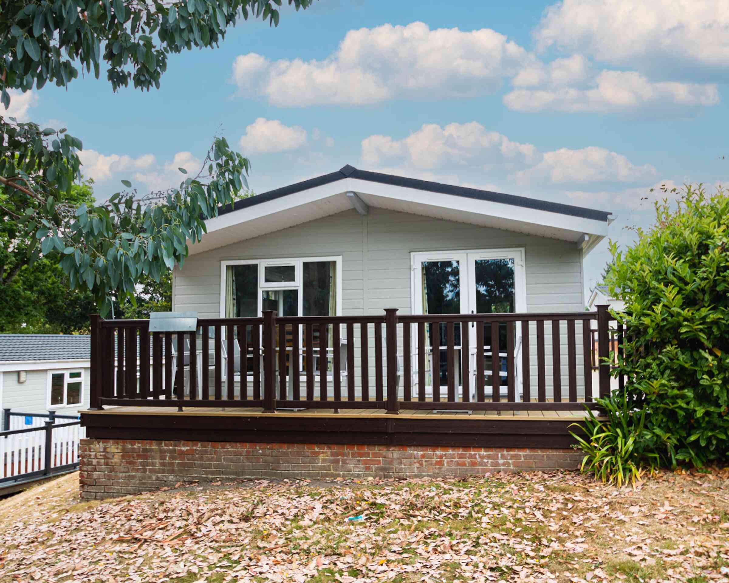 A modern single-story house with a sloped roof, featuring a wooden deck surrounded by greenery and pastel blue skies with fluffy clouds in the background.