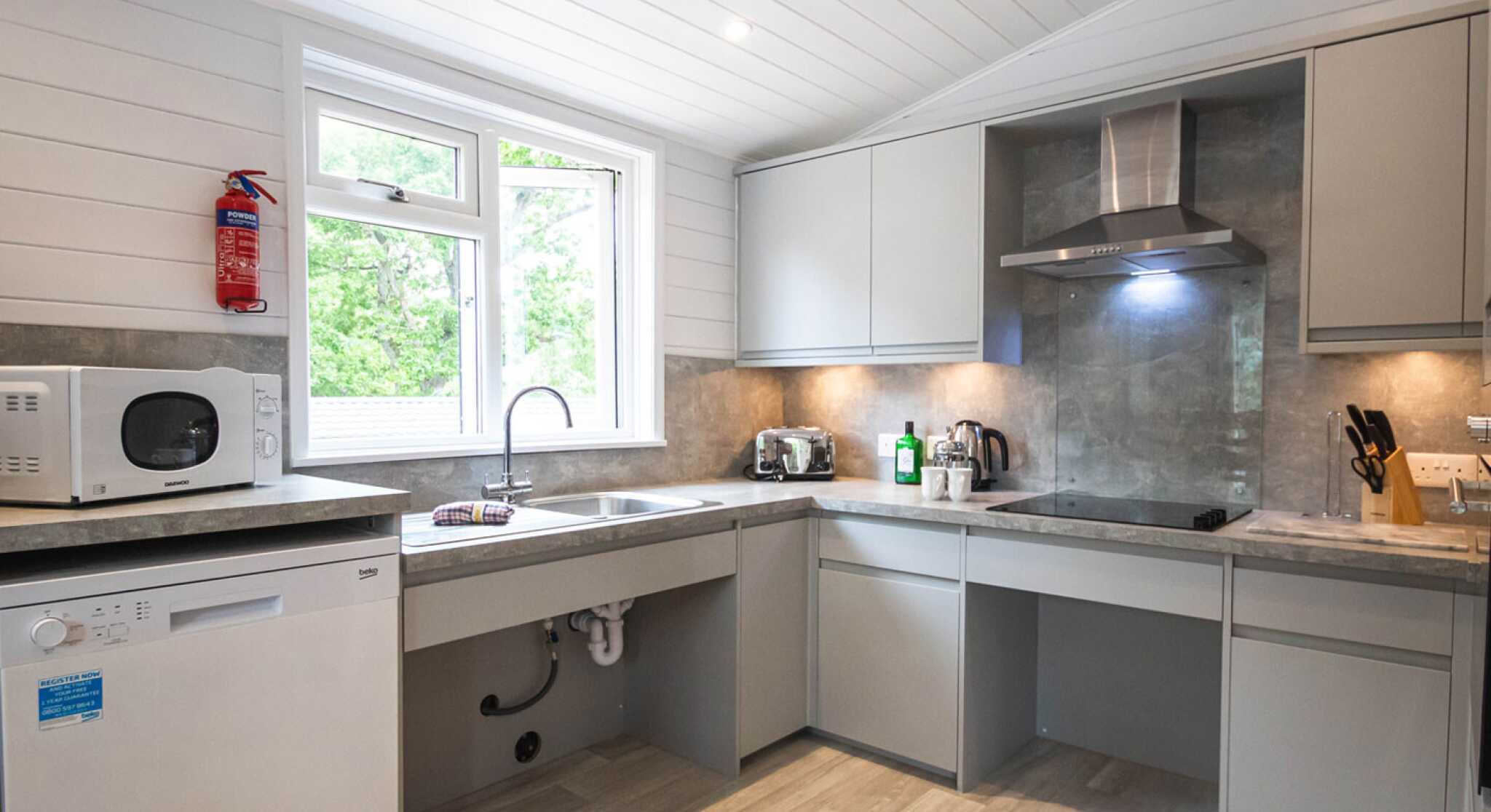 Modern kitchen with light gray cabinets, a stainless steel microwave, and a dishwasher. A large window provides natural light, and there are small appliances on the countertops. The flooring is light in color, enhancing the airy feel of the space.