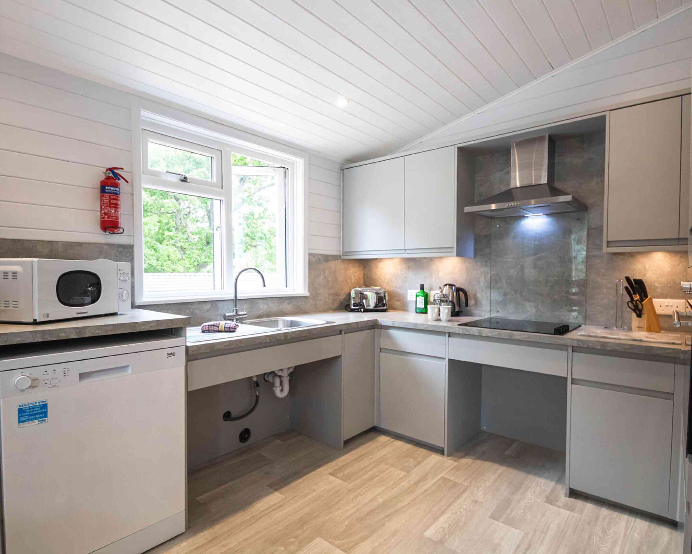 Modern kitchen with light gray cabinets, a stainless steel microwave, and a dishwasher. A large window provides natural light, and there are small appliances on the countertops. The flooring is light in color, enhancing the airy feel of the space.