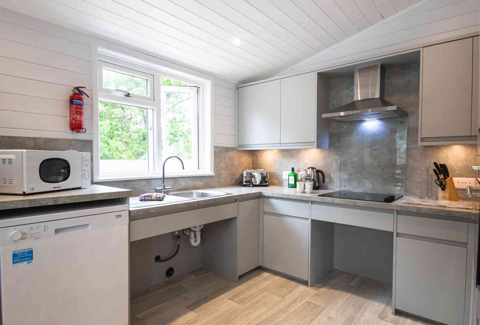 Modern kitchen with light gray cabinets, a stainless steel microwave, and a dishwasher. A large window provides natural light, and there are small appliances on the countertops. The flooring is light in color, enhancing the airy feel of the space.