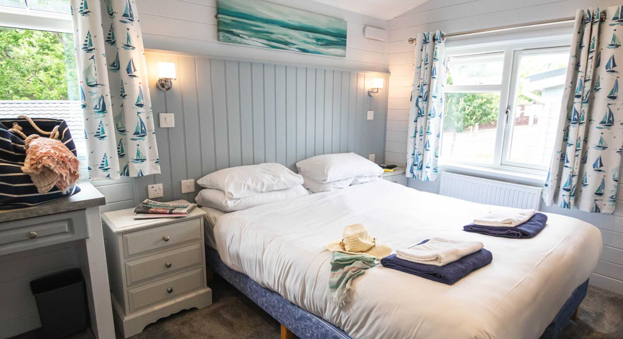 A cozy bedroom with two neatly made beds, gray walls, and nautical-themed curtains. A small side table holds a decorative shell, and a painting of the ocean hangs above the bed. Light streams in from a window, creating a bright and inviting atmosphere.