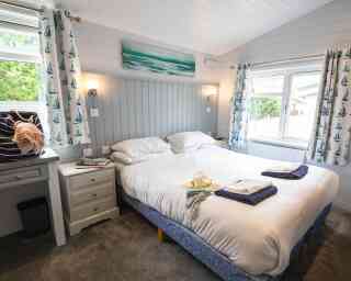 A cozy bedroom with two neatly made beds, gray walls, and nautical-themed curtains. A small side table holds a decorative shell, and a painting of the ocean hangs above the bed. Light streams in from a window, creating a bright and inviting atmosphere.