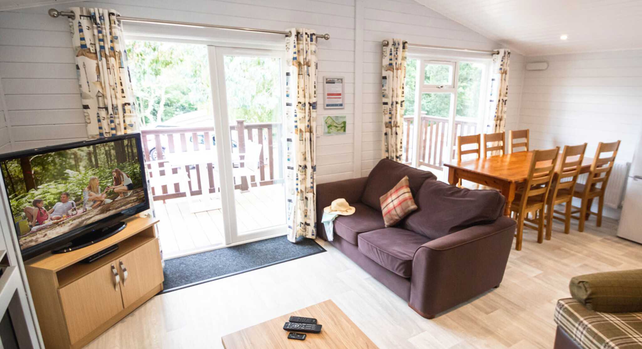 Cozy living area with a large sofa, coffee table, and TV, leading to a deck through sliding glass doors. A dining table with wooden chairs is visible in the background. Natural light filters in through windows, enhancing the inviting atmosphere.