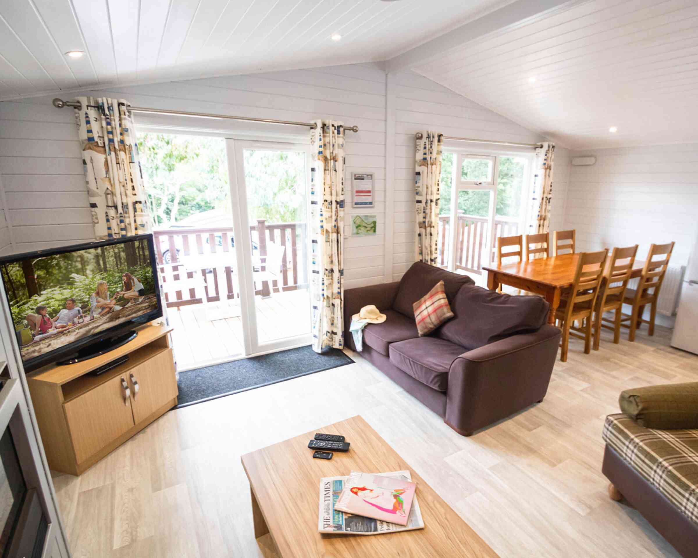 Cozy living area with a large sofa, coffee table, and TV, leading to a deck through sliding glass doors. A dining table with wooden chairs is visible in the background. Natural light filters in through windows, enhancing the inviting atmosphere.