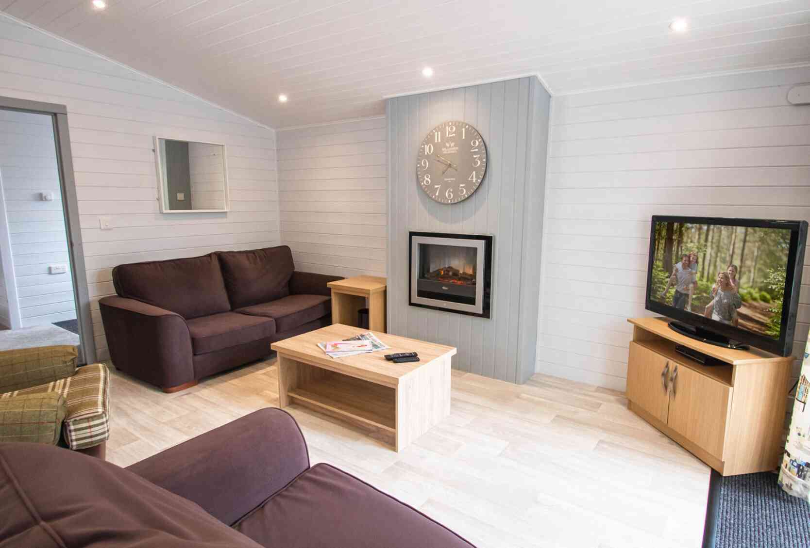 A cozy living room featuring a dark brown sofa, a light wood coffee table, and a wooden TV cabinet with a television displaying a scene. A digital clock hangs on the wall above a modern electric fireplace. The room has light-colored walls and bright lighting.