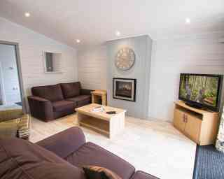 A cozy living room featuring a dark brown sofa, a light wood coffee table, and a wooden TV cabinet with a television displaying a scene. A digital clock hangs on the wall above a modern electric fireplace. The room has light-colored walls and bright lighting.