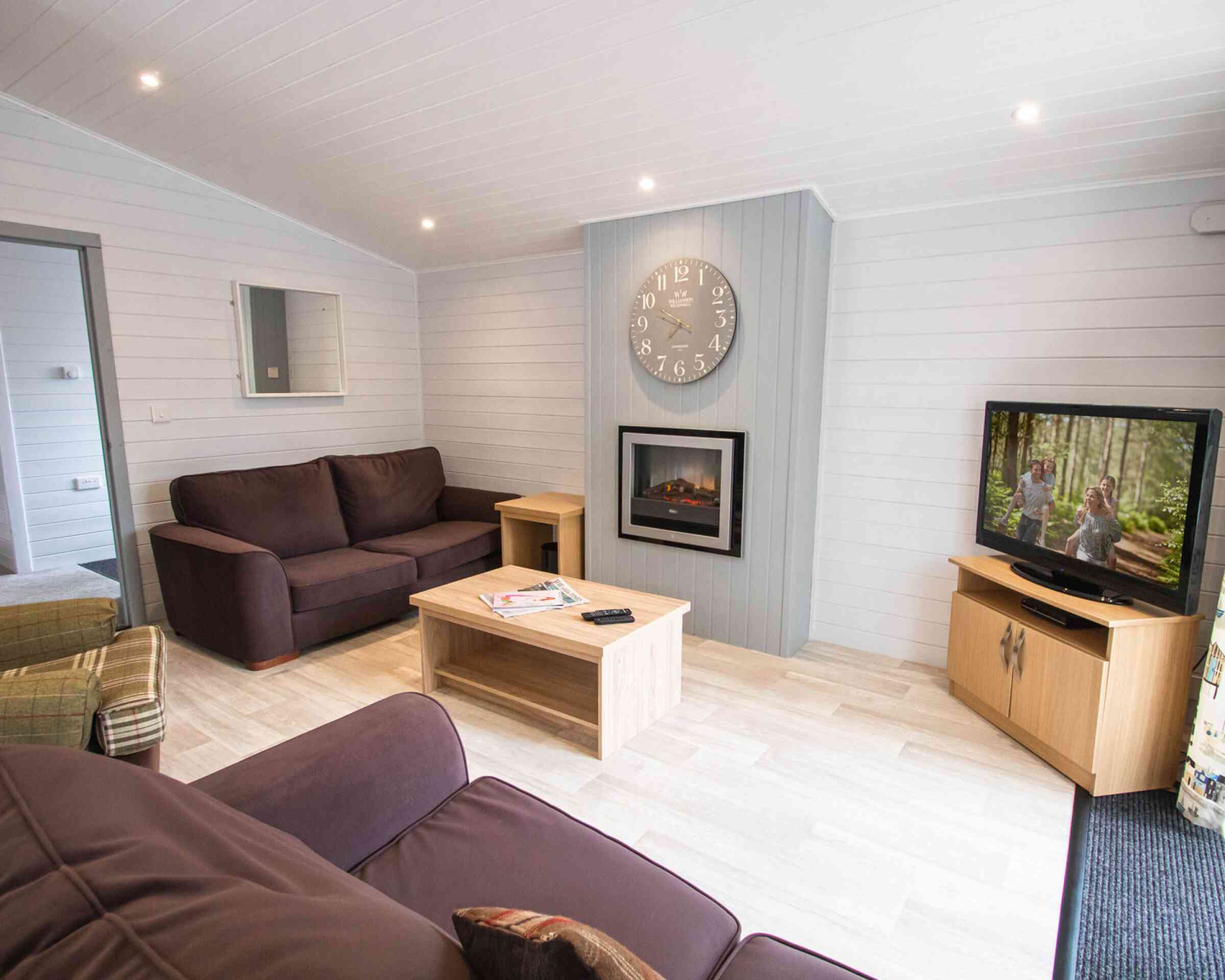 A cozy living room featuring a dark brown sofa, a light wood coffee table, and a wooden TV cabinet with a television displaying a scene. A digital clock hangs on the wall above a modern electric fireplace. The room has light-colored walls and bright lighting.