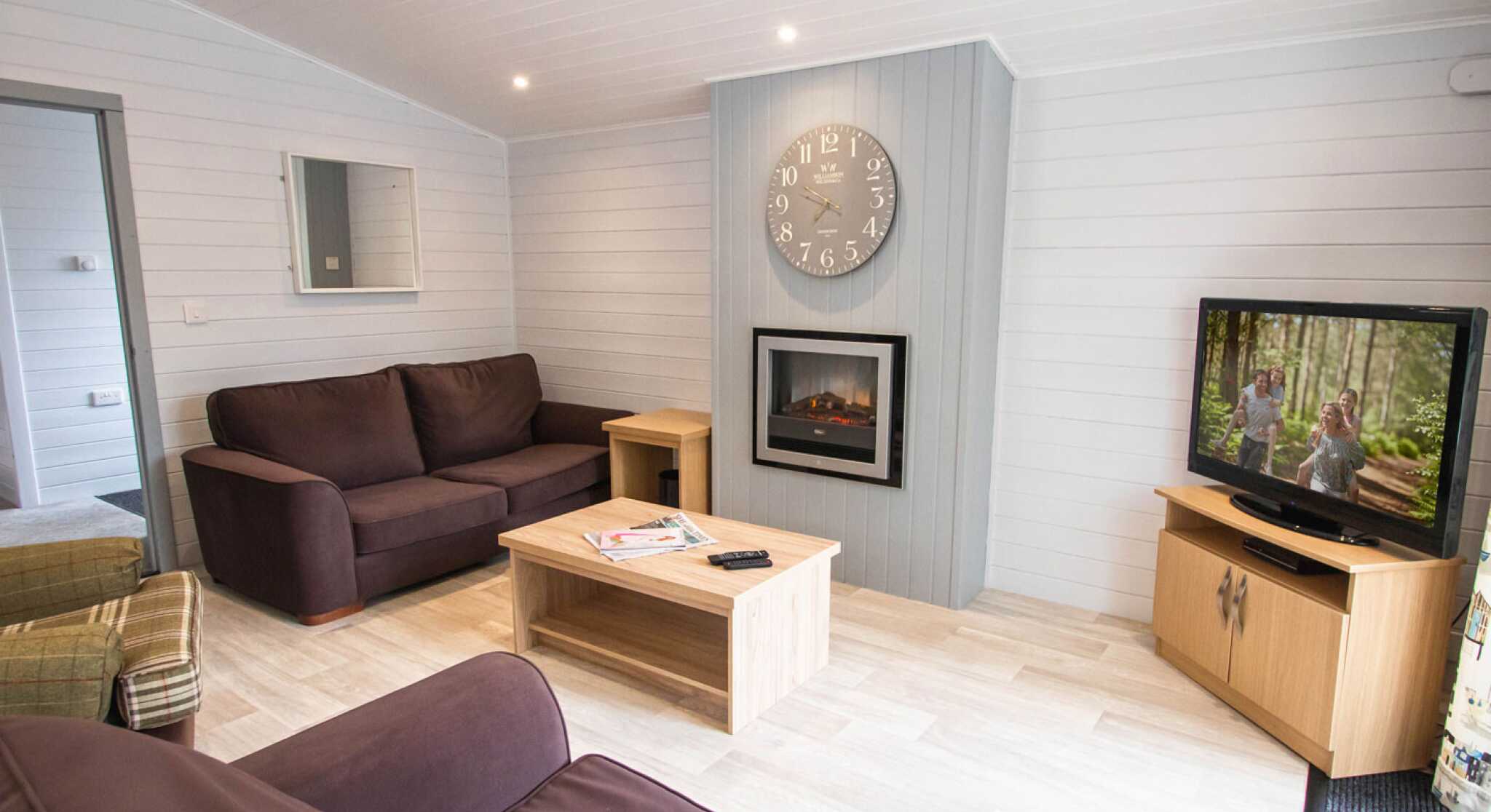 A cozy living room featuring a dark brown sofa, a light wood coffee table, and a wooden TV cabinet with a television displaying a scene. A digital clock hangs on the wall above a modern electric fireplace. The room has light-colored walls and bright lighting.