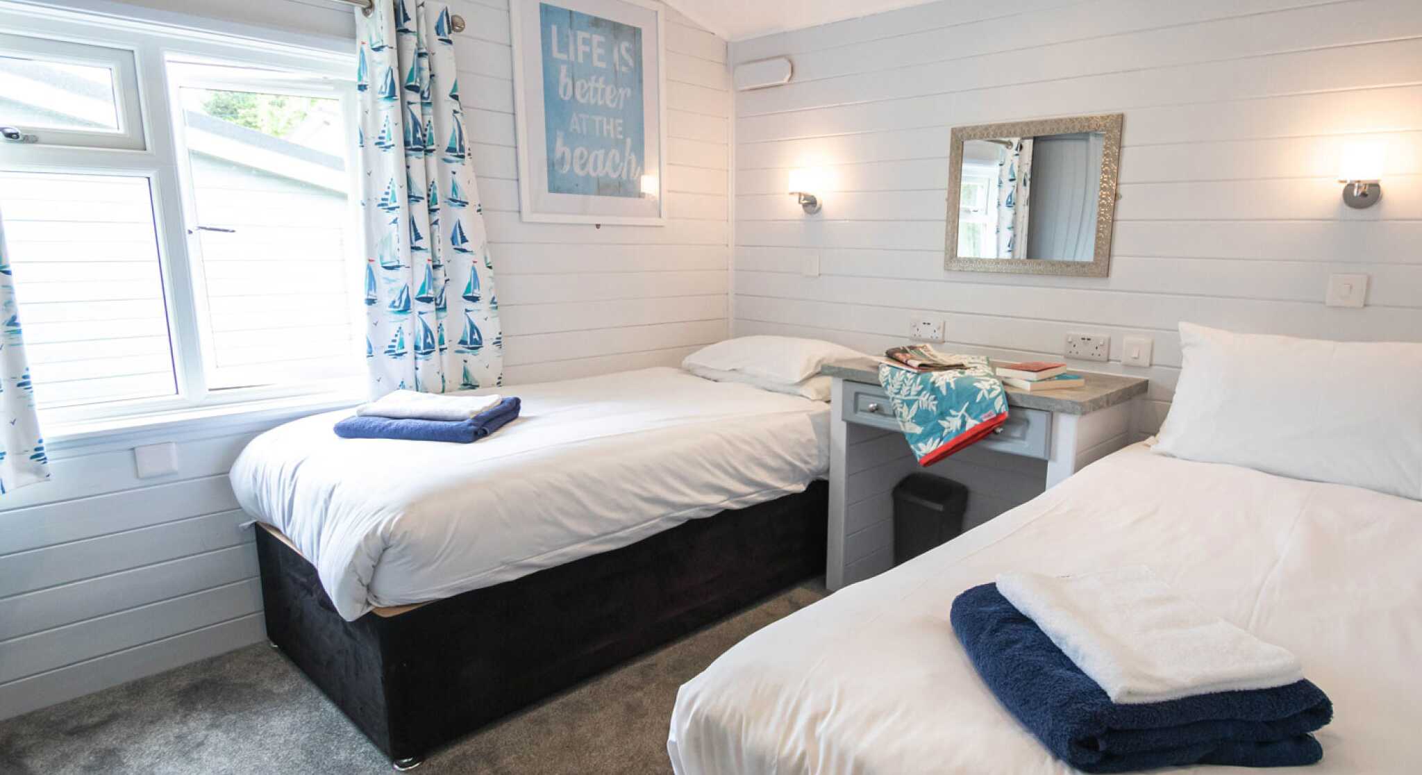 A cozy bedroom featuring two single beds with folded towels on each. A window with nautical-themed curtains allows natural light to enter. A mirror and small desk are visible, along with a motivational beach-themed artwork on the wall. The carpeted floor adds warmth to the space.