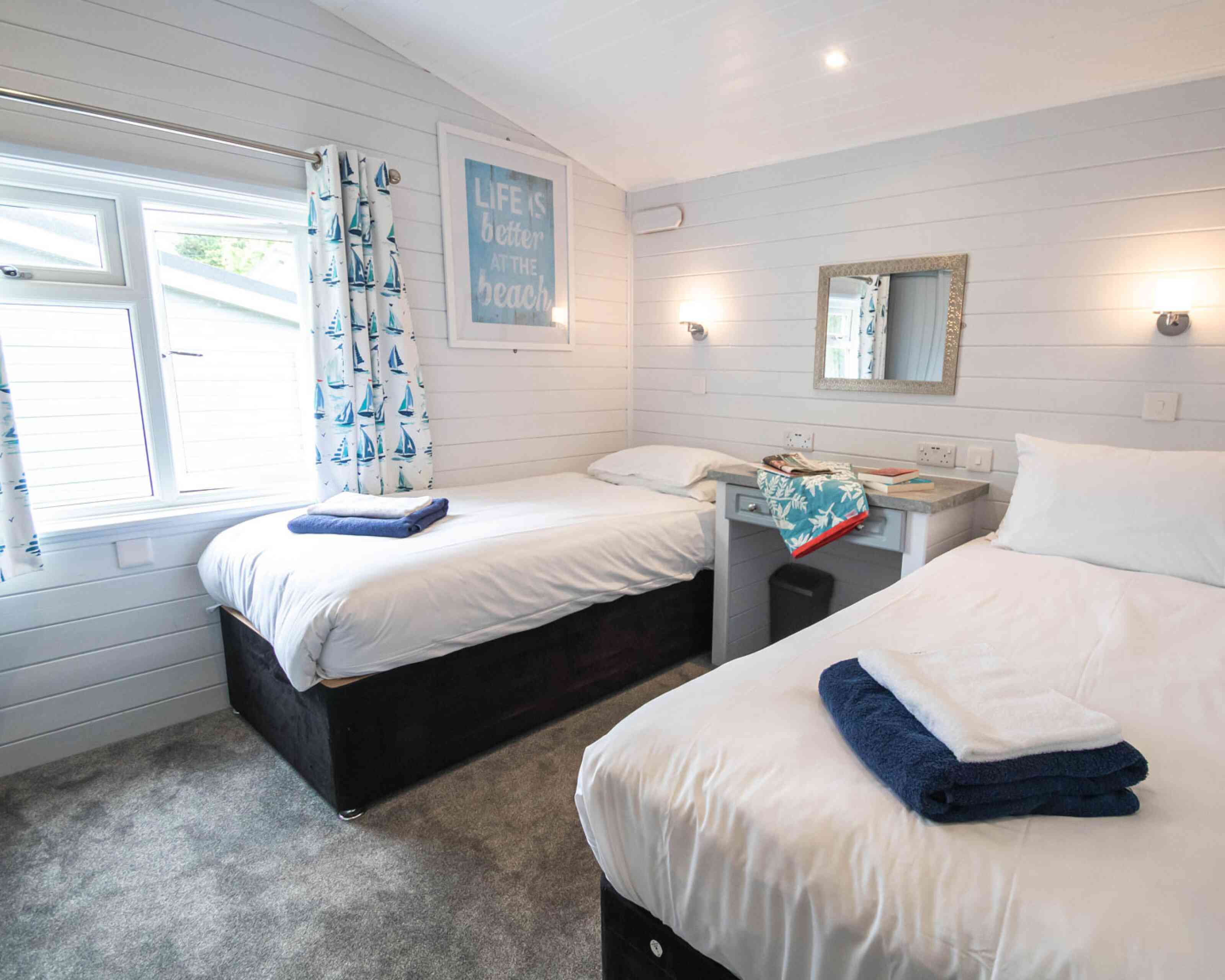 A cozy bedroom featuring two single beds with folded towels on each. A window with nautical-themed curtains allows natural light to enter. A mirror and small desk are visible, along with a motivational beach-themed artwork on the wall. The carpeted floor adds warmth to the space.