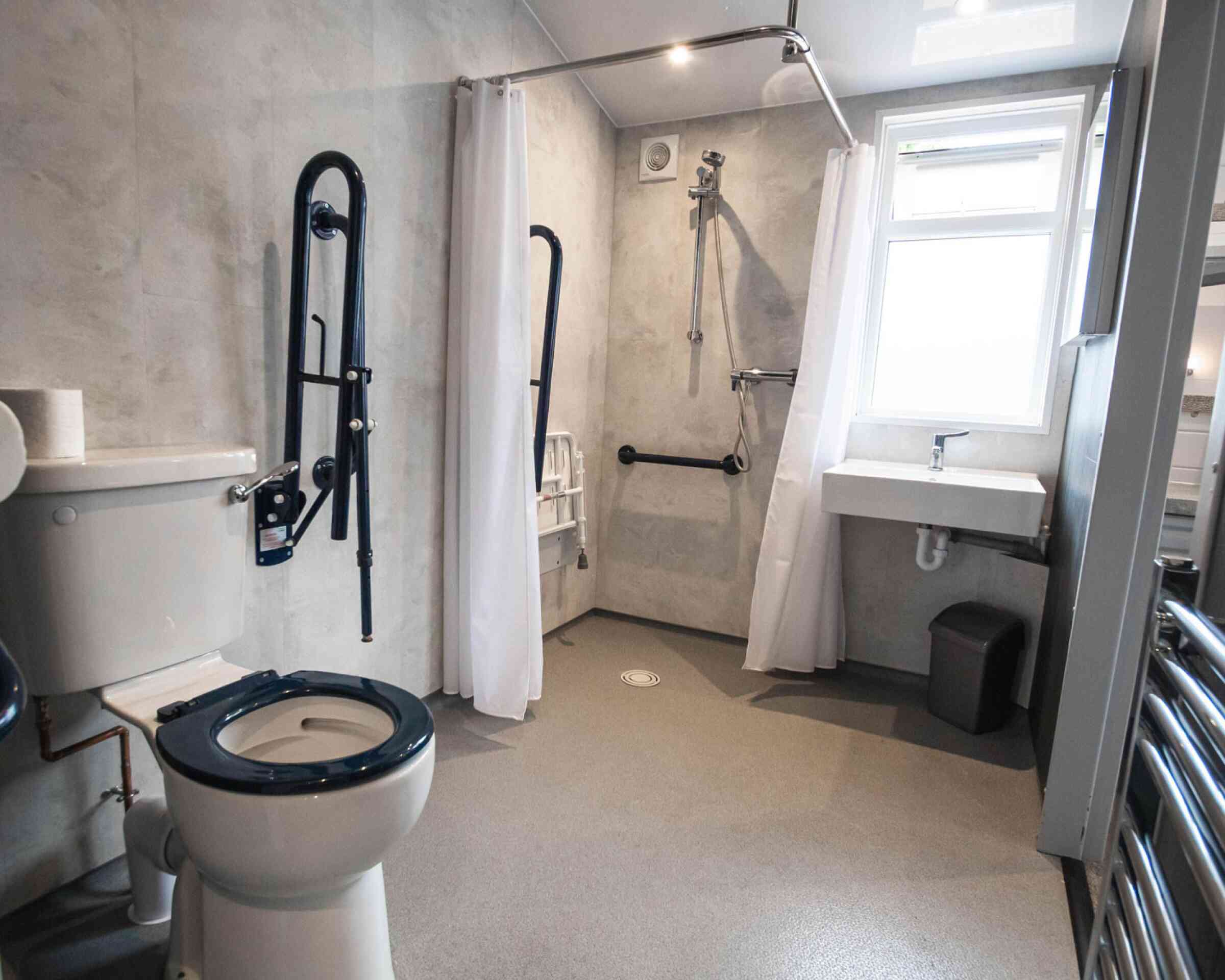 A bathroom with a toilet and accessible shower area. The walls are textured grey, featuring grab bars and a shower chair. A small sink is adjacent to the shower, and there is a window providing natural light.