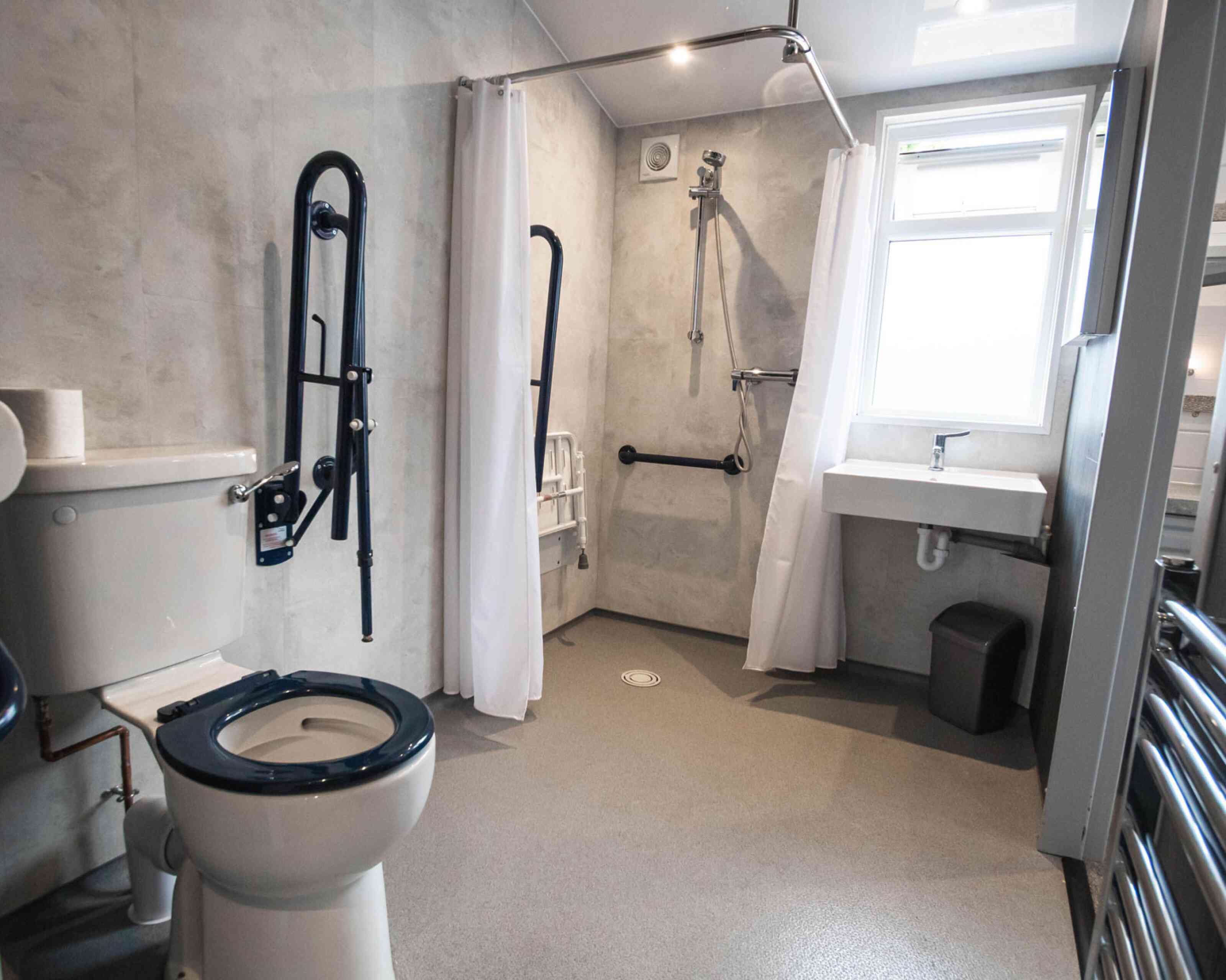 A bathroom with a toilet and accessible shower area. The walls are textured grey, featuring grab bars and a shower chair. A small sink is adjacent to the shower, and there is a window providing natural light.