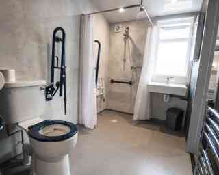 A bathroom with a toilet and accessible shower area. The walls are textured grey, featuring grab bars and a shower chair. A small sink is adjacent to the shower, and there is a window providing natural light.