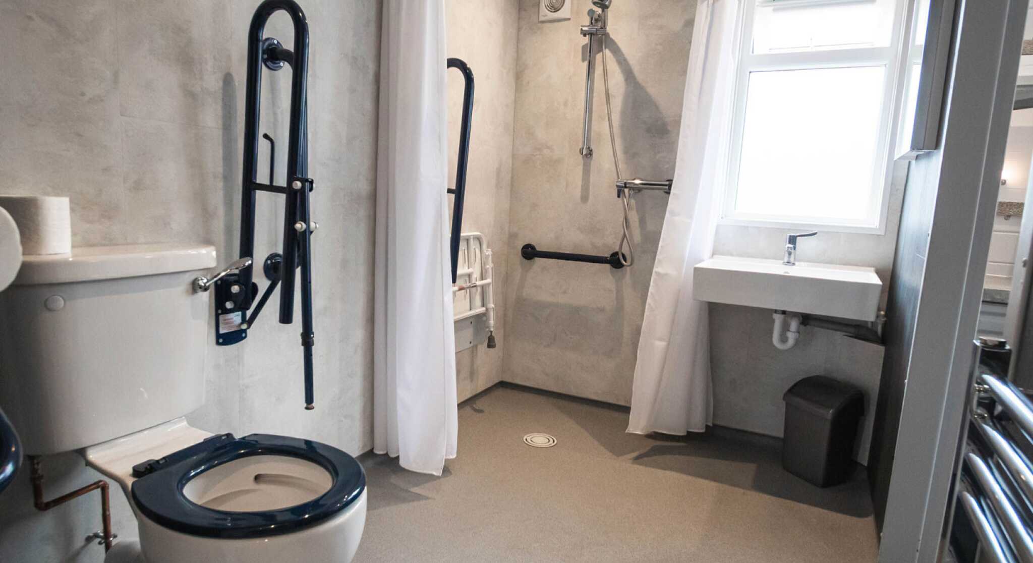 A bathroom with a toilet and accessible shower area. The walls are textured grey, featuring grab bars and a shower chair. A small sink is adjacent to the shower, and there is a window providing natural light.