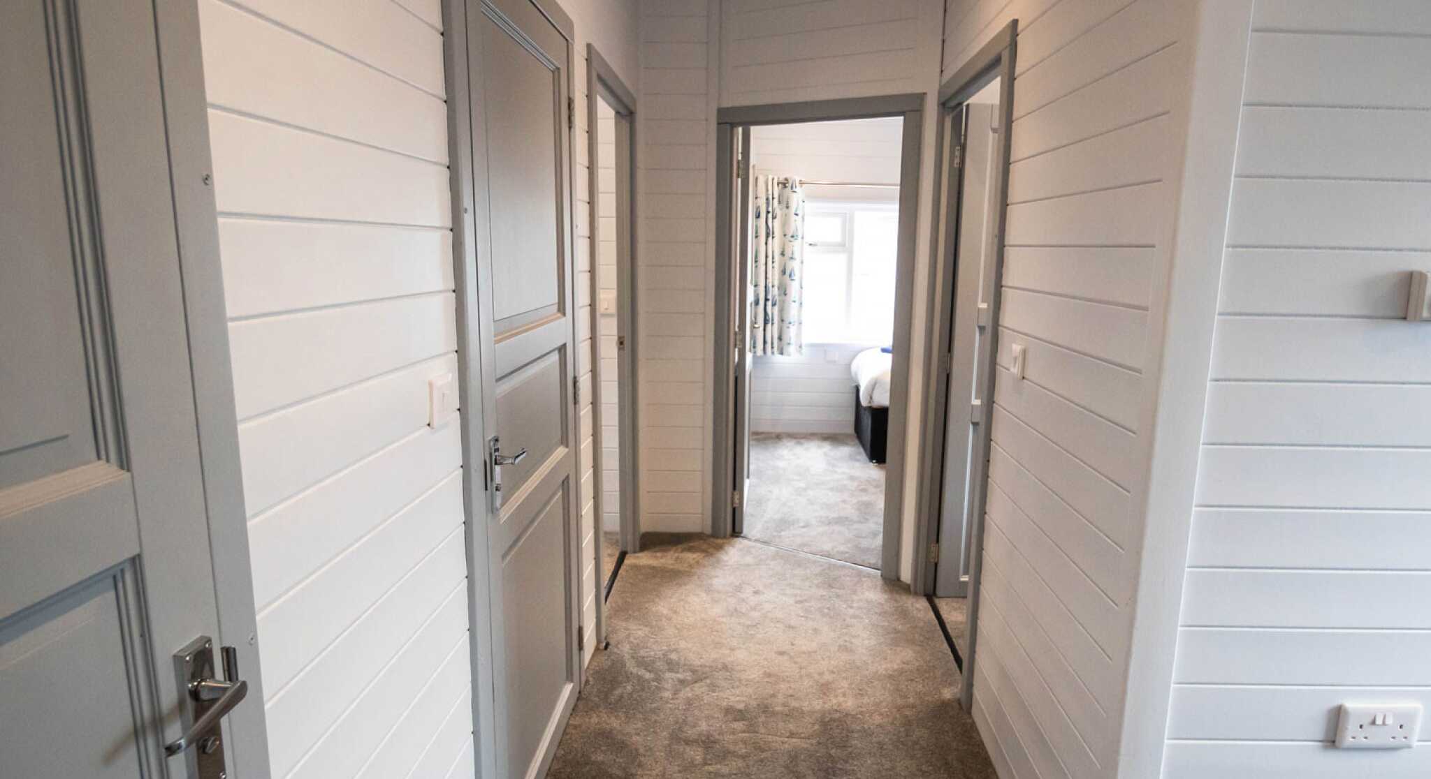 A well-lit hallway featuring light gray paneling and beige carpet. There are three doorways leading to different rooms, with a glimpse of a bedroom at the end. The walls are white with a clean and modern aesthetic.