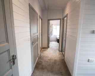 A well-lit hallway featuring light gray paneling and beige carpet. There are three doorways leading to different rooms, with a glimpse of a bedroom at the end. The walls are white with a clean and modern aesthetic.